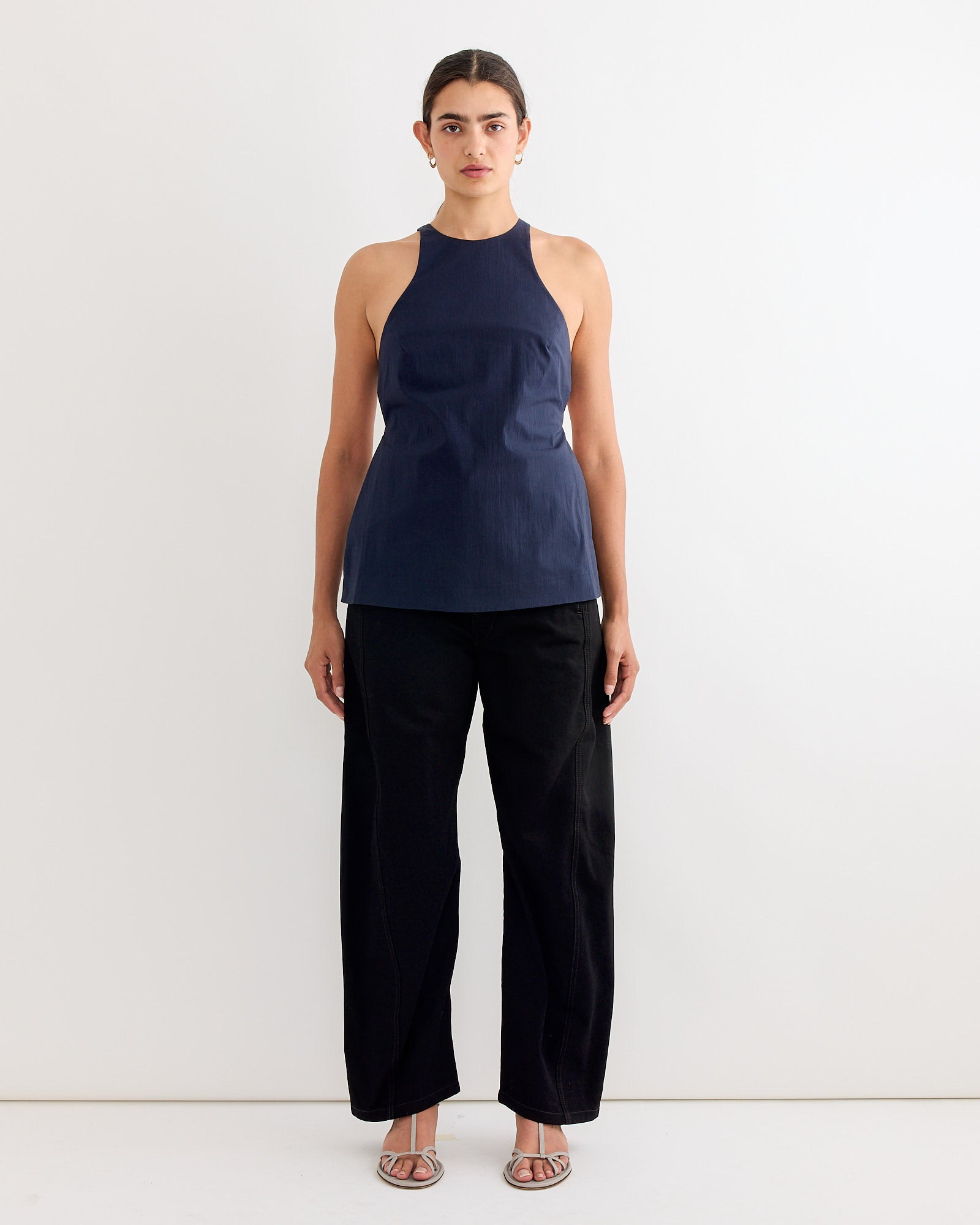 A woman with dark hair pulled back and a neutral expression poses against a white background, wearing the ST. AGNI Racerback Top in Dark Ink, loose black pants, and open-toed sandals.