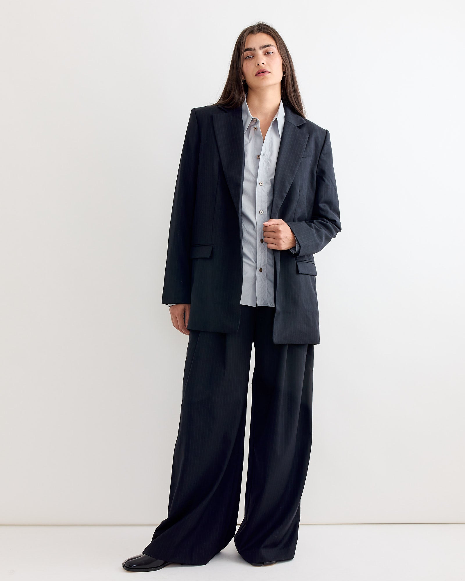A person with long dark hair stands against a white wall, confidently wearing an oversized ST. AGNI Split Back Blazer in Black Pinstripe, paired with wide-leg trousers and a light blue button-up shirt for a modern suiting look.