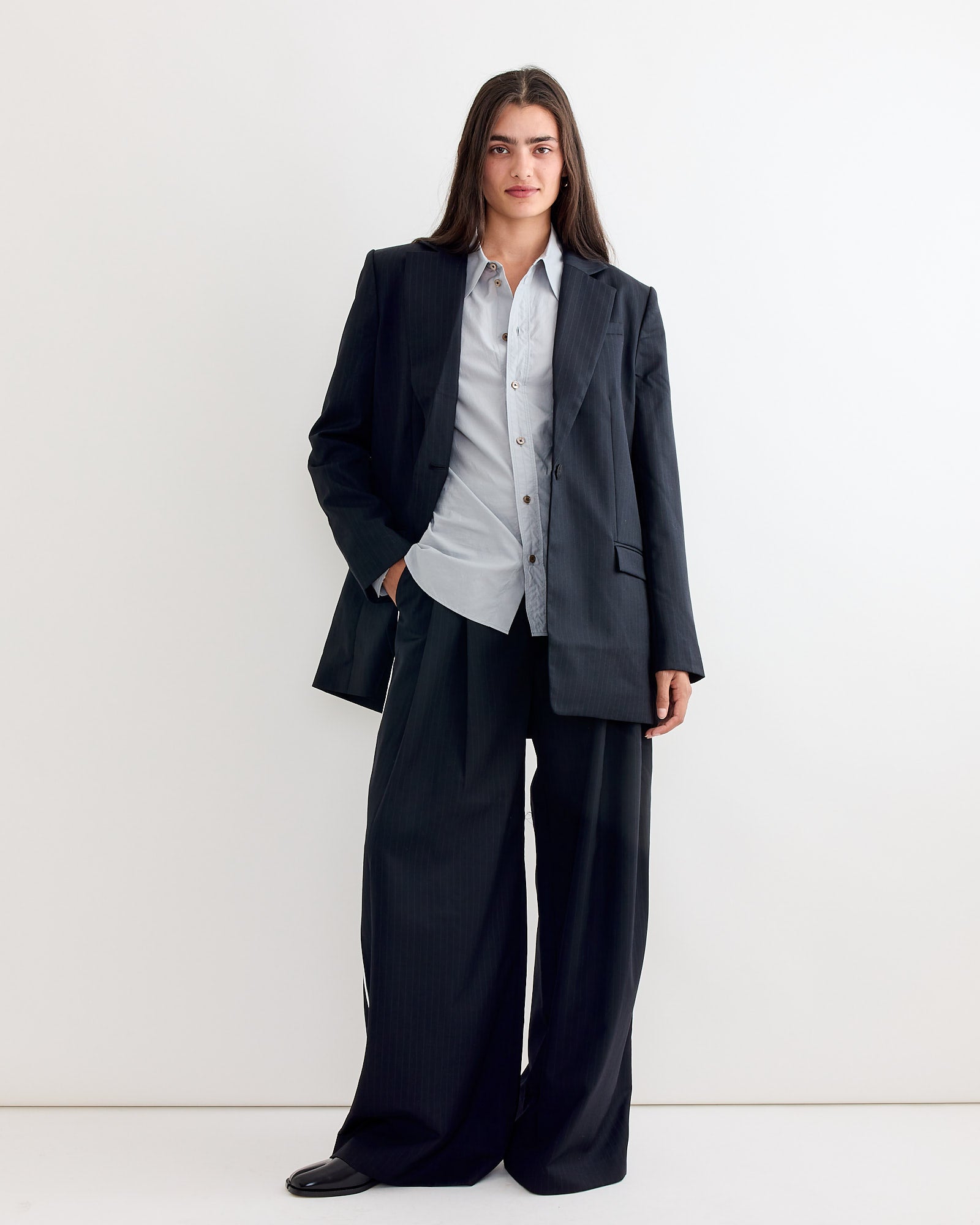 A person with long dark hair stands against a white background wearing the ST. AGNI Side Stripe Homme Pant in Black Pinstripe, paired with a matching oversized jacket and light blue shirt for a relaxed, versatile look.