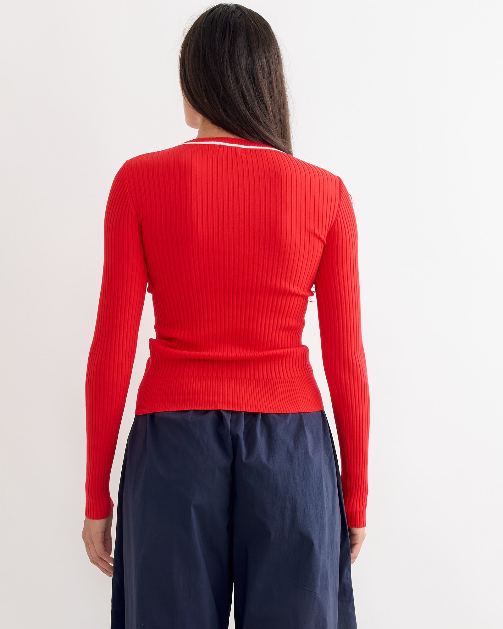 A person with long dark hair is seen from the back wearing the ST. AGNI Fine Line Top in Red/Off White with a ribbed crew neckline and loose navy blue pants, against a plain white background.