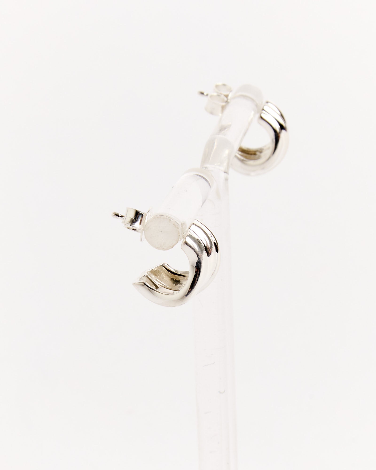 The Recycled Silver Earrings by Completedworks are small, shiny hoops shown on a clear acrylic stand against a white background. For sizing or fit questions, our online chat team is ready to help.