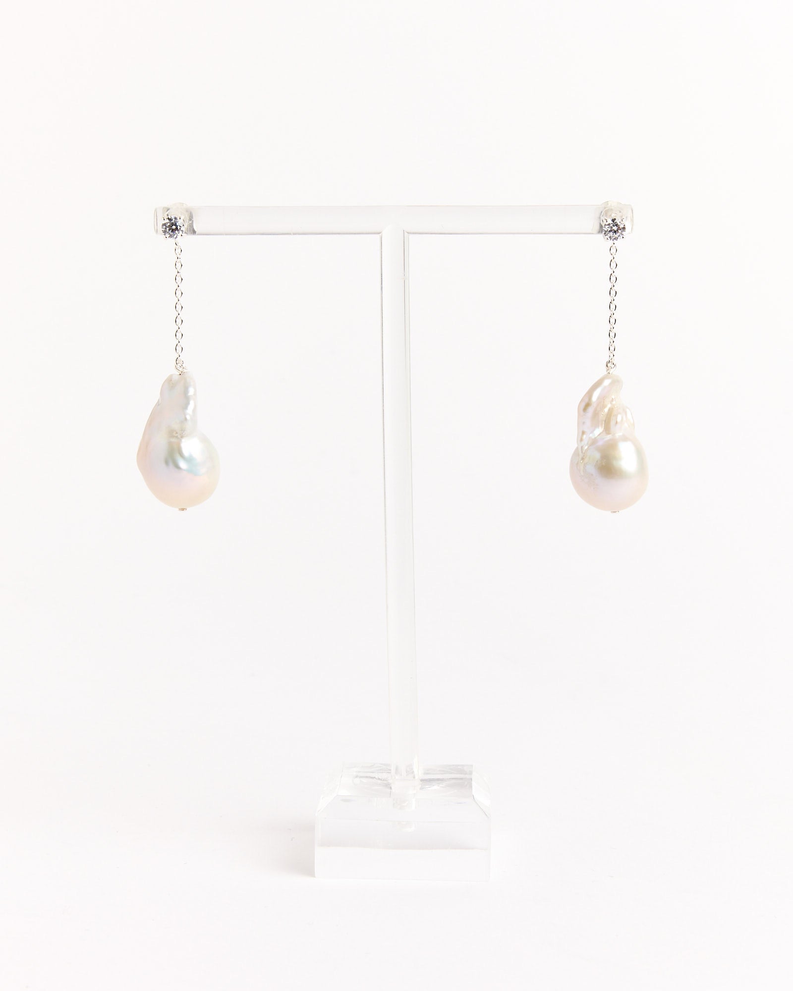 Recycled Silver Baroque Pearl CZ Earrings by Completedworks feature irregular white pearls on delicate chains, displayed on a clear T-stand against a white background.