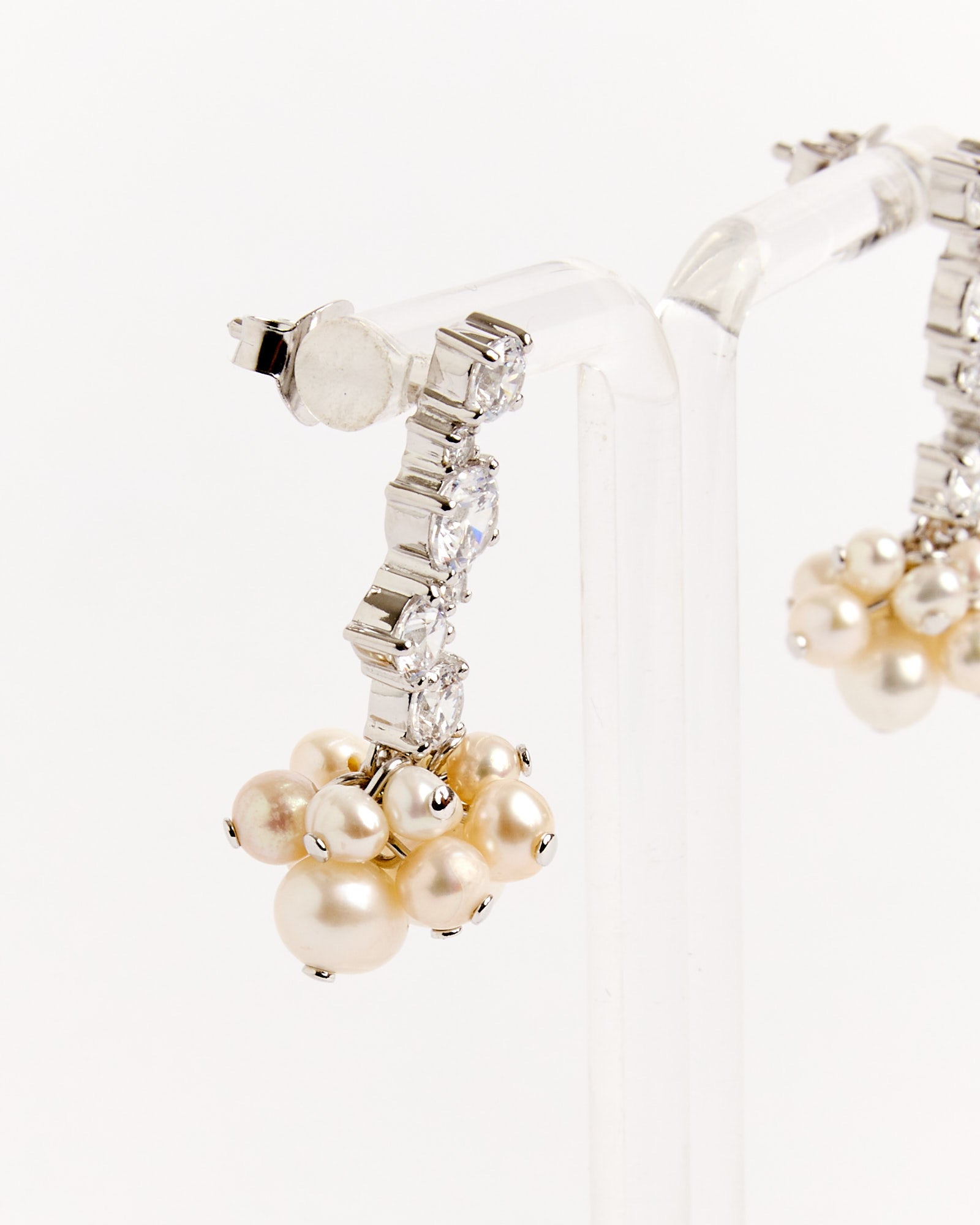 A close-up of Completedworks Recycled Silver Pearl Cluster CZ Earrings, featuring clear gemstones and cream-colored pearls, displayed on a transparent stand. For sizing or fit questions, contact our online chat support.