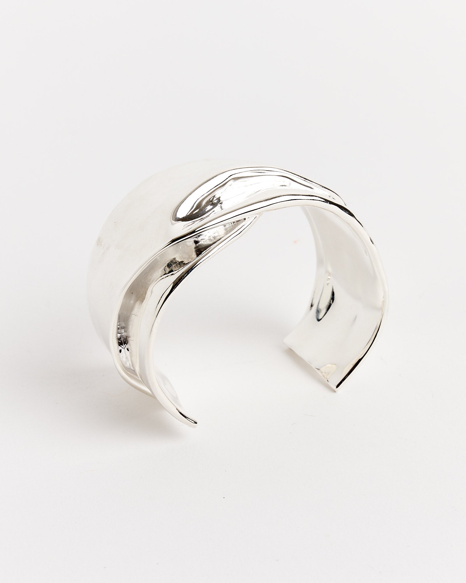 The Completedworks Small Brass Cuff, featuring a fluid, wavy design with matte and shiny finishes, sits on a plain white background. This open-ended bracelet ensures a comfortable fit for most wrists.