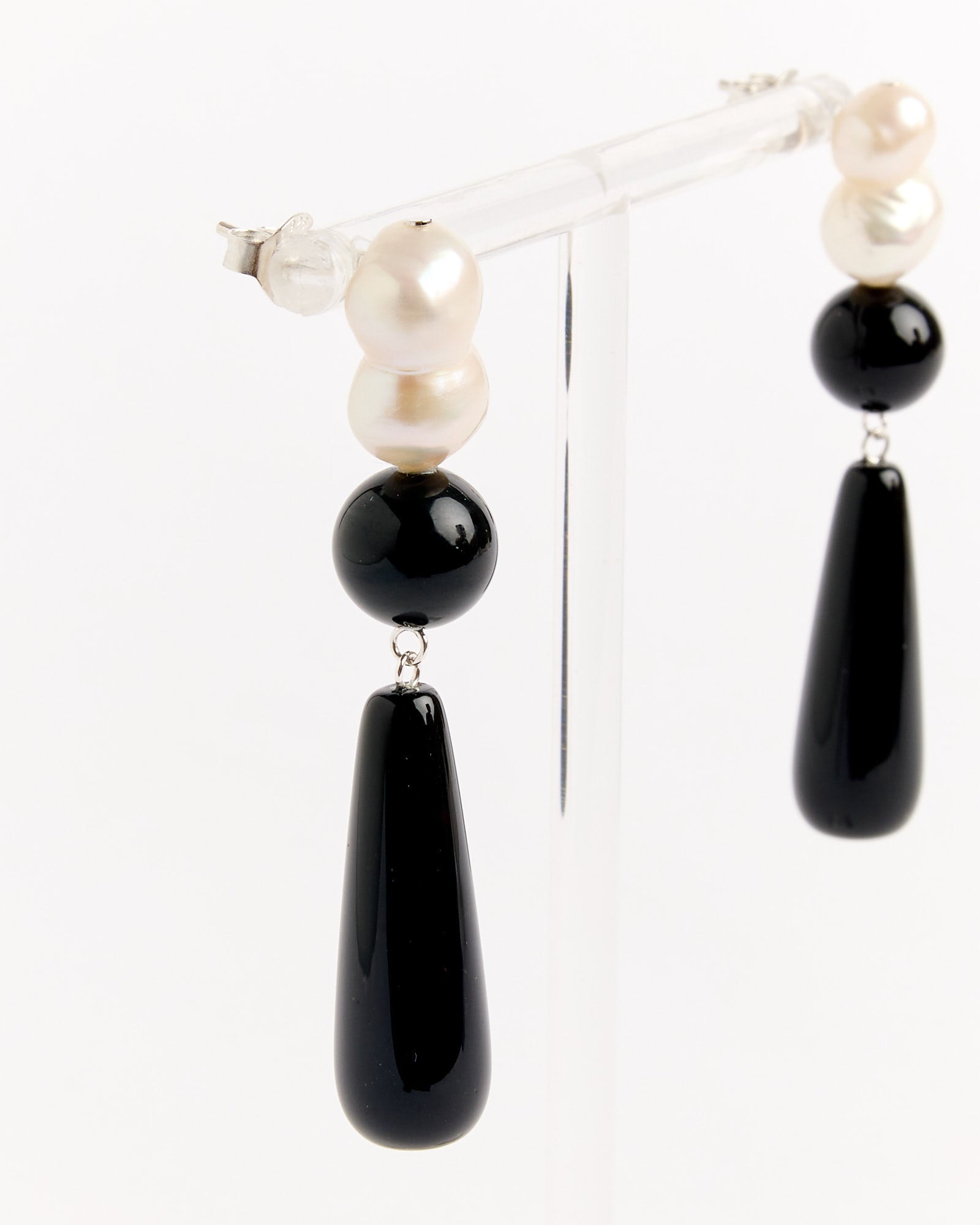 The Completedworks Recycled Silver Black Onyx FWP Earrings feature two white round pearls, a round black onyx bead, and a teardrop-shaped black pendant. Shown on a clear stand with a white background. Chat with us online for sizing and fit help.