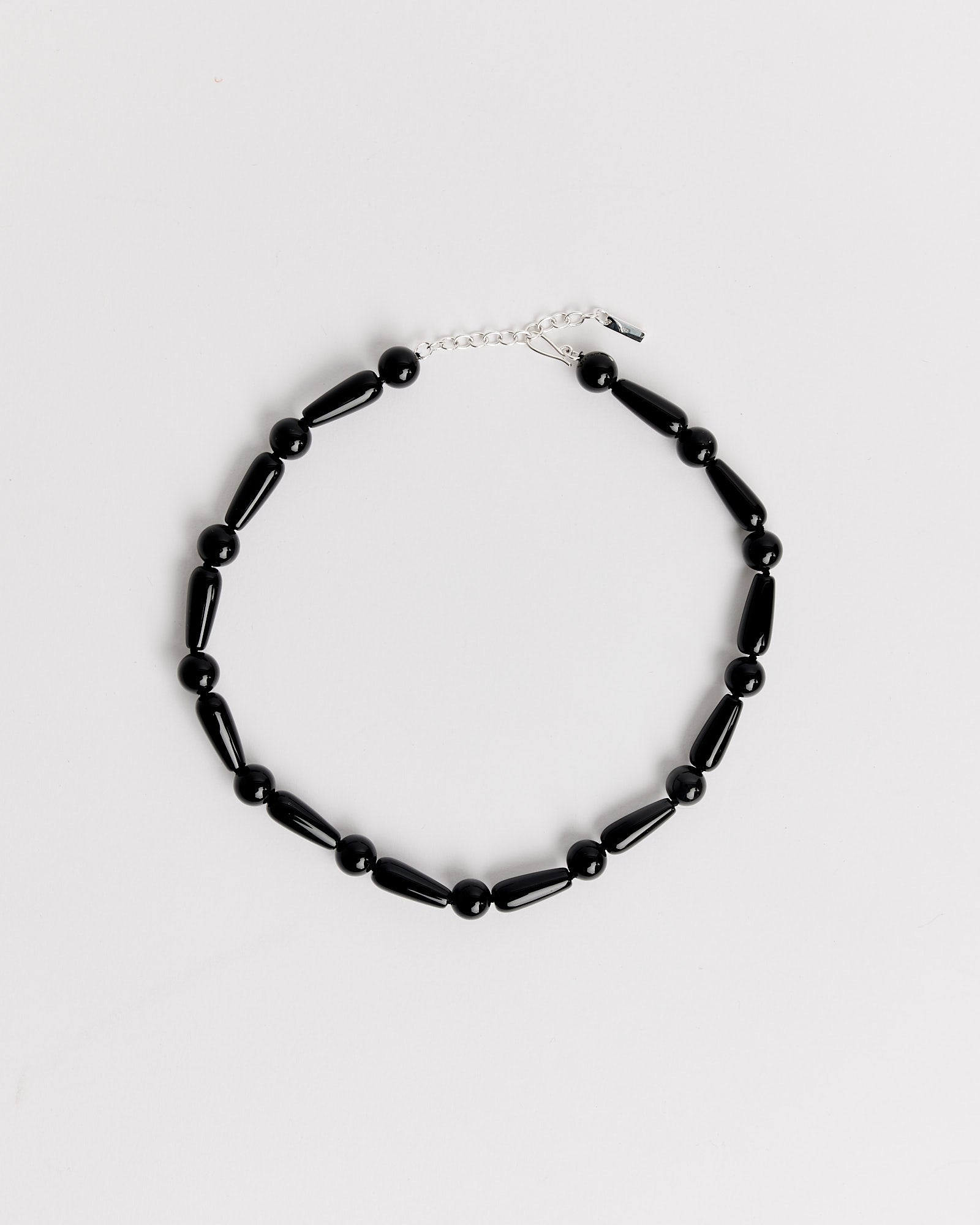 The Depths Of Time Necklace in Black Onyx/Silver Plated by Completedworks features alternating round and cylindrical black onyx beads, a silver clasp with an adjustable chain, and flexible sizing, all arranged on a plain white background.
