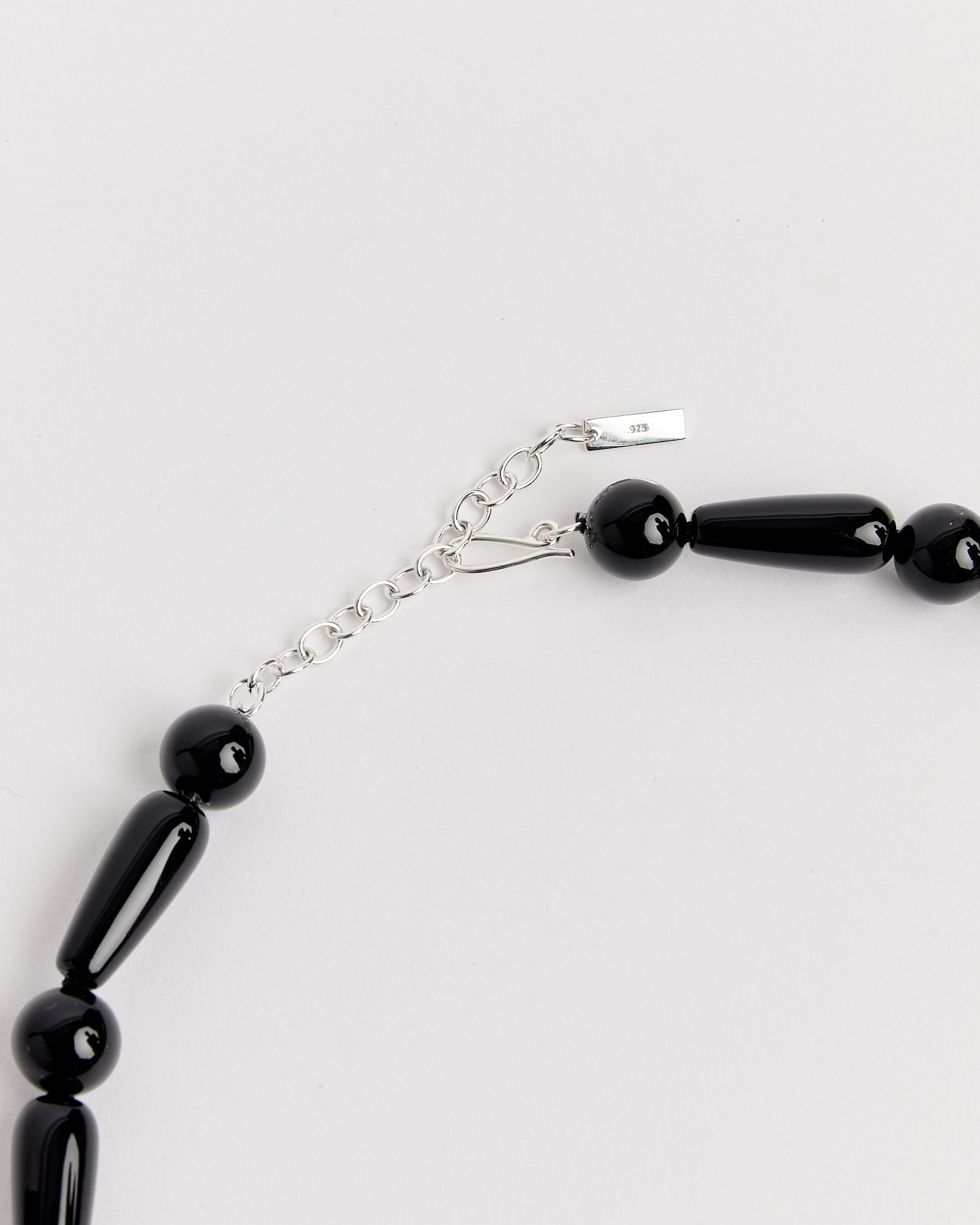 A close-up of Completedworks' The Depths Of Time Necklace in Black Onyx/Silver Plated, showcasing elongated and round black beads with a silver chain extender and clasp—ideal for adjustable sizing and comfortable wear.