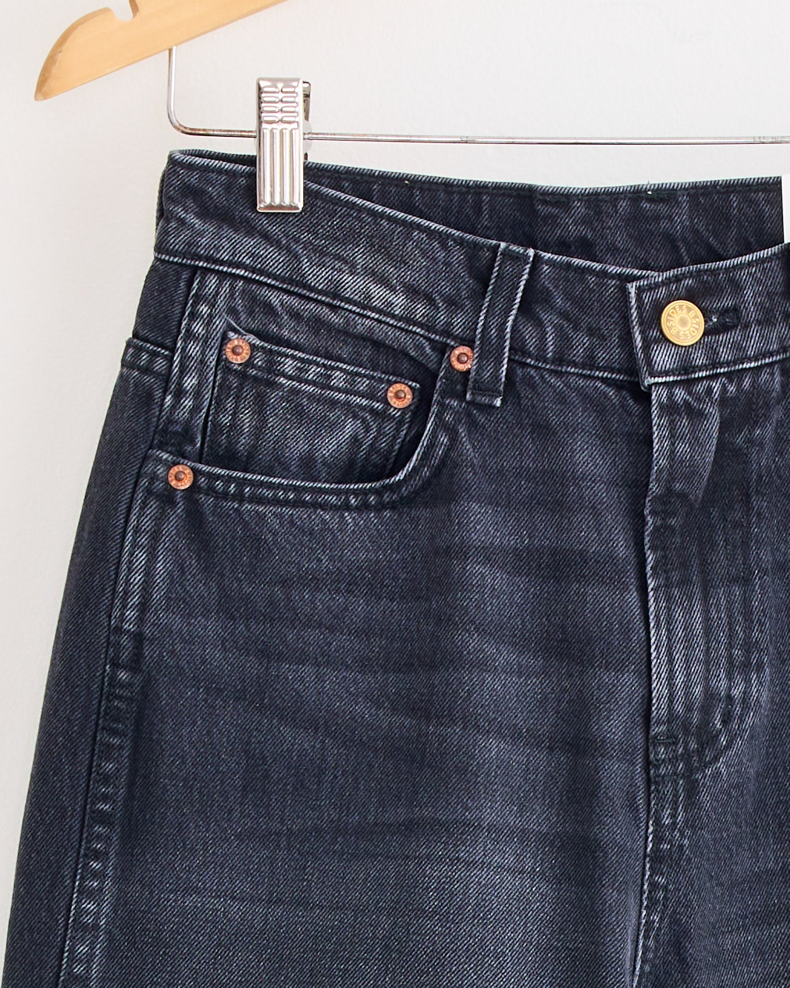 A close-up of the Plein Jean in Stil Black by B Sides, hanging on a wooden hanger, highlights the front pocket, coin pocket, belt loops, copper rivets, and metallic waistband button.
