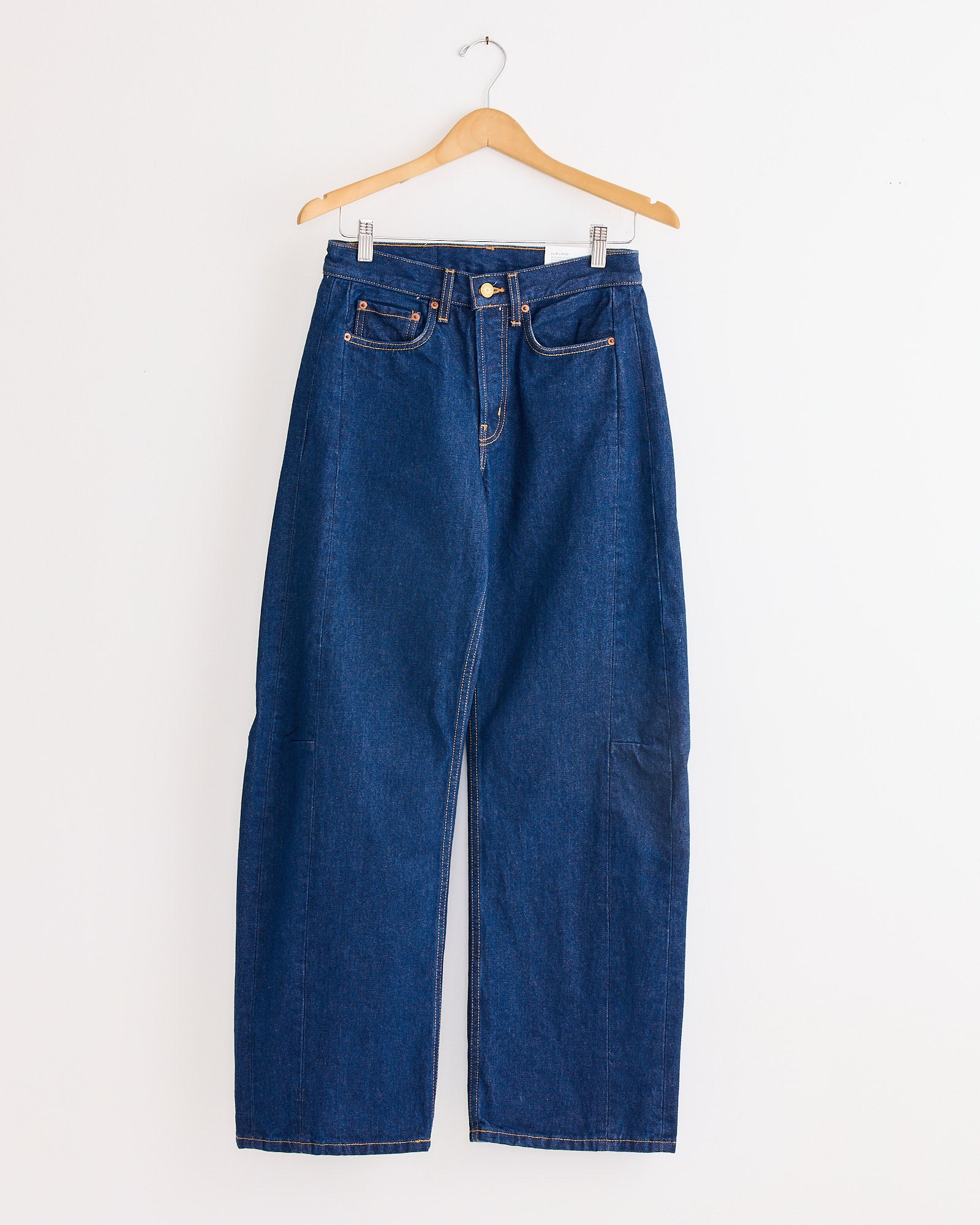 The Slim Lasso Jean in Indigo Rinse by B Sides hangs on a wooden hanger against a plain white wall, showcasing its high waist, wide-leg silhouette, front and back pockets, visible stitching, and belt loops.