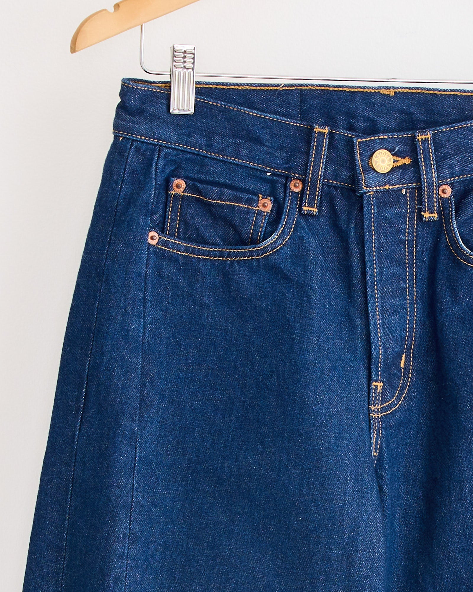 A close-up of the Slim Lasso Jean in Indigo Rinse by B Sides, hanging on a wooden hanger, highlights the front pockets, button, zipper, and distinctive orange stitching details.