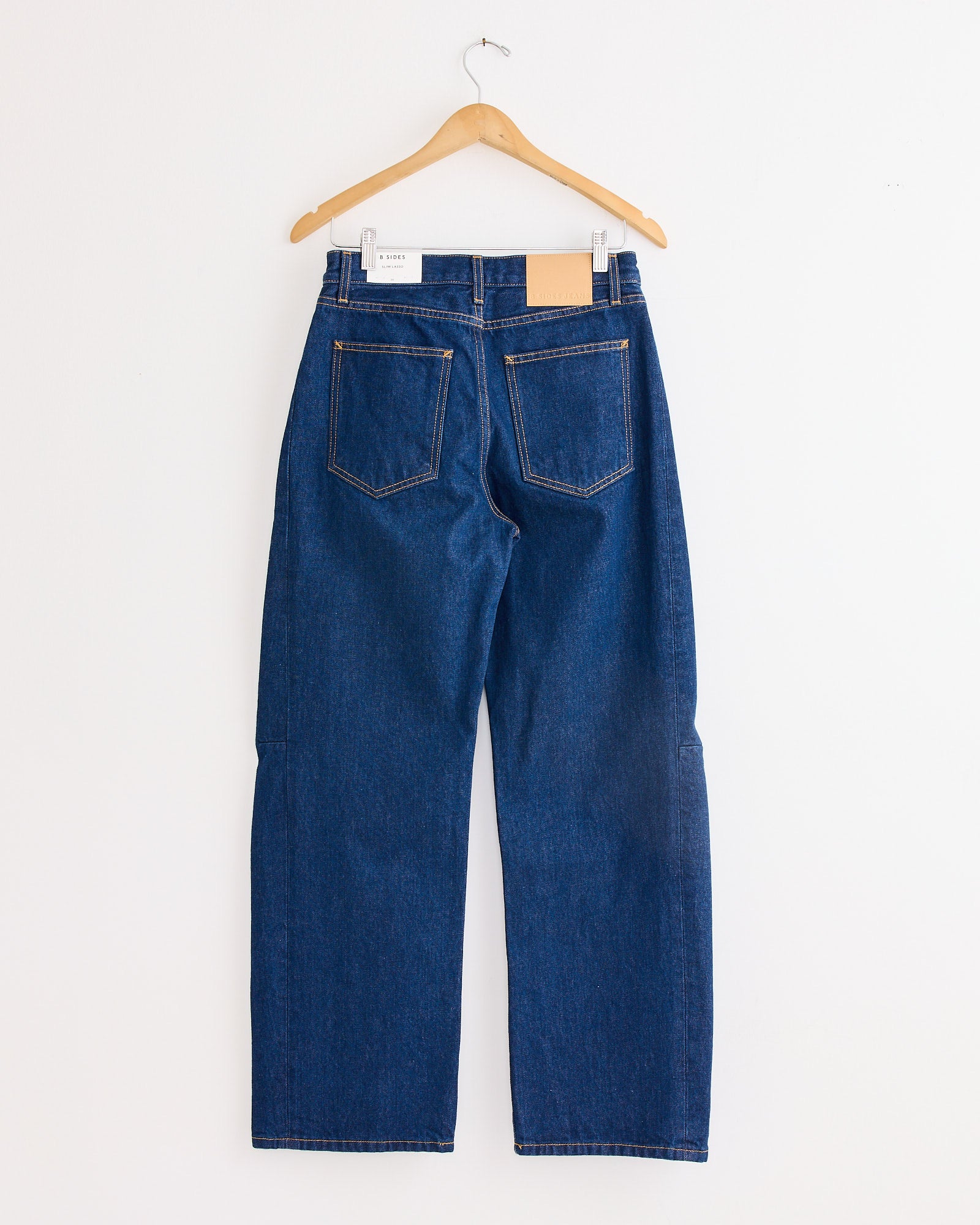 The Slim Lasso Jean in Indigo Rinse by B Sides, featuring two back pockets, hangs on a wooden hanger against a plain white wall.