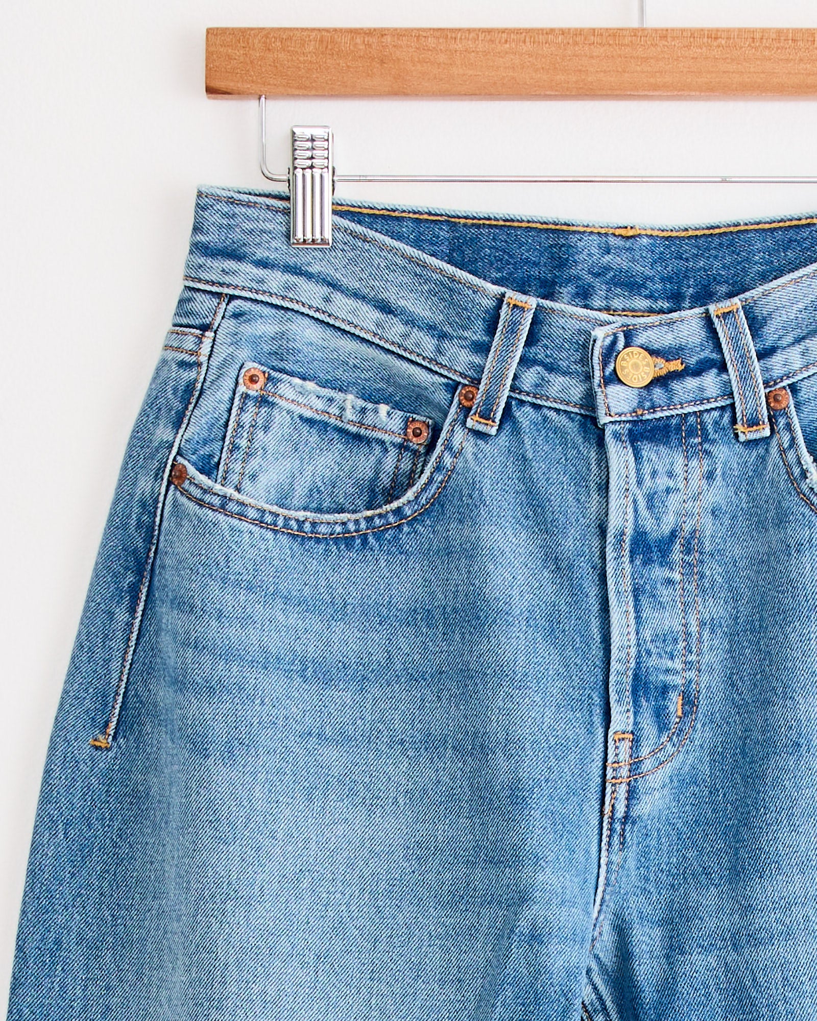 A close-up of the Tilda Jean in Jane Wash by B Sides, a light blue indigo denim mid-rise straight style, hanging on a wooden hanger and highlighting the waistband, front button, zipper, pockets, and belt loops.