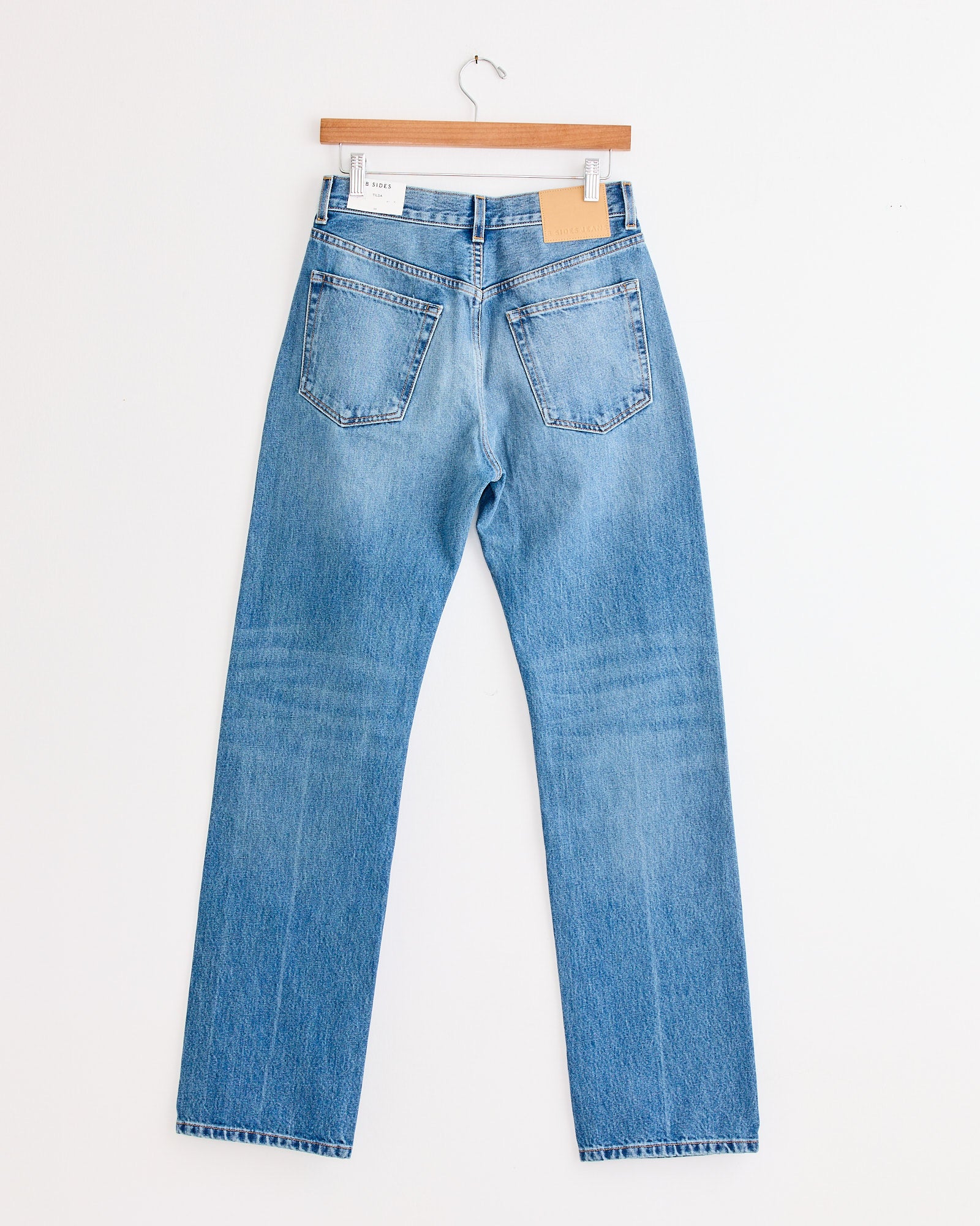 The B Sides Tilda Jean in Jane Wash, a mid-rise straight-leg light blue denim, hangs on a wooden hanger against a white wall, displayed from the back showing two rear pockets.