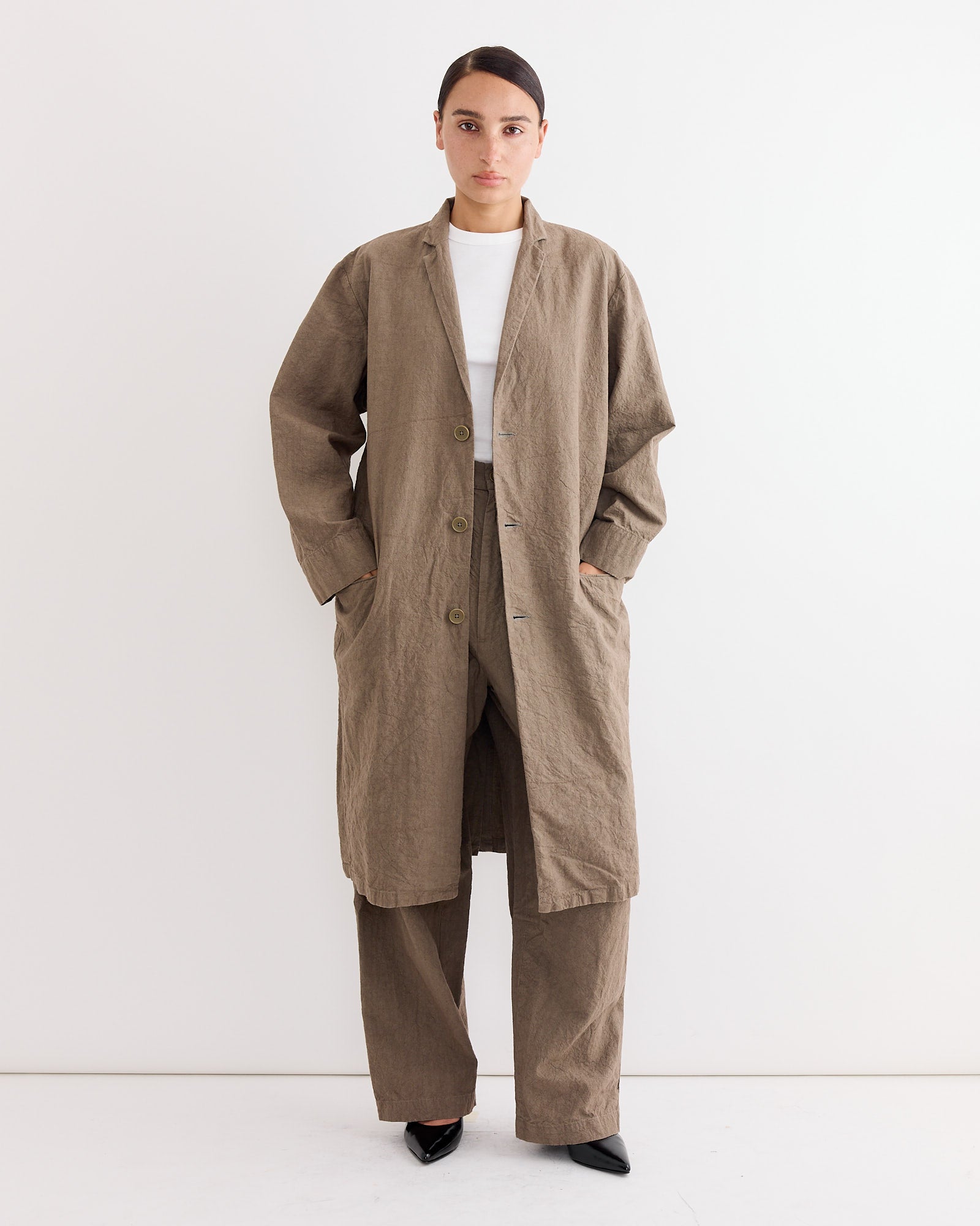 A person stands against a plain white wall, wearing Black Crane’s Crinkled Trouser in Khaki with a long linen coat, a white top, and black pointed shoes, hands in the coat pockets.