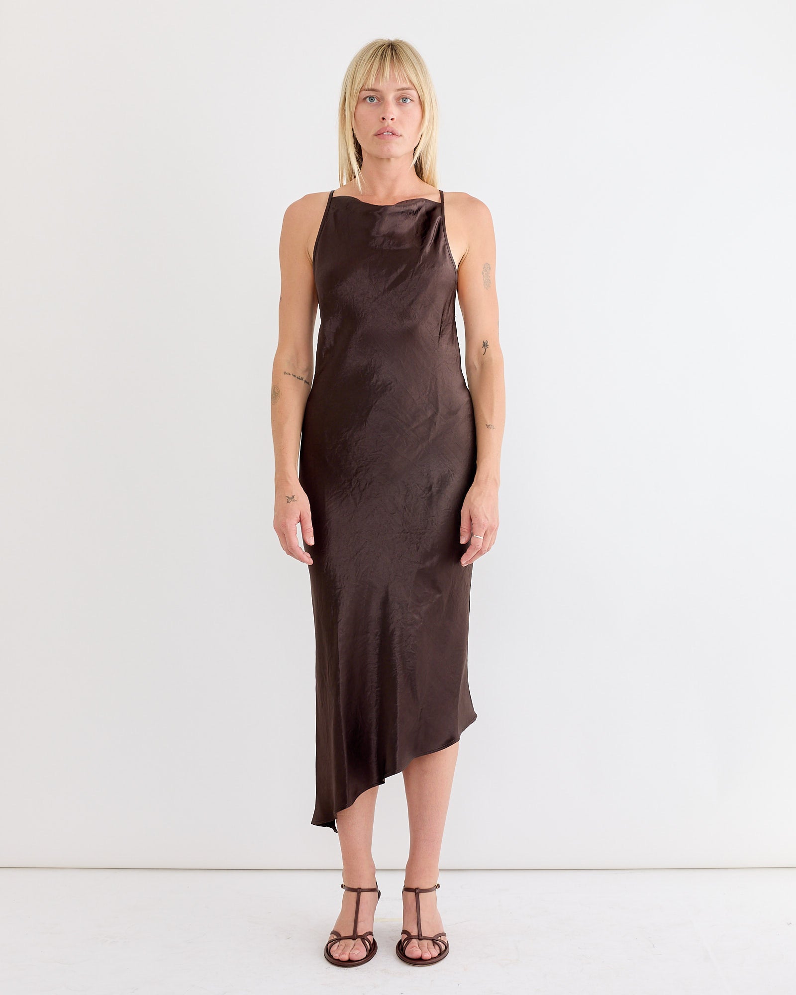 A woman with straight blonde hair wears the NOMIA Crossback Bias Dress in Dark Brown, featuring adjustable spaghetti straps and an asymmetrical hem, styled with brown strappy sandals against a plain white background.