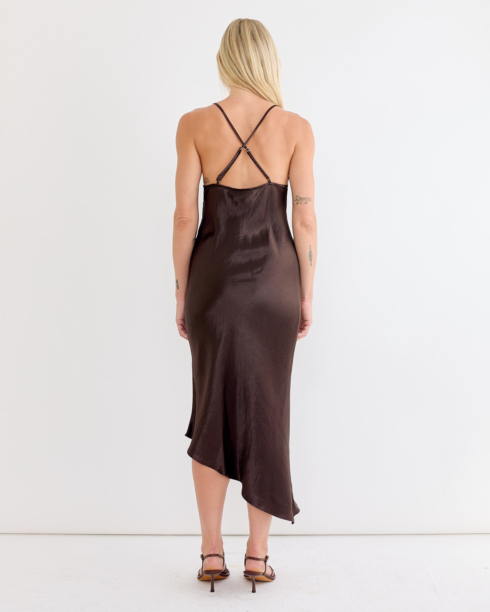 A woman with long blonde hair wears the NOMIA Crossback Bias Dress in Dark Brown, featuring silky Japanese washed satin and adjustable spaghetti straps, standing facing away in a minimal, white-walled room.