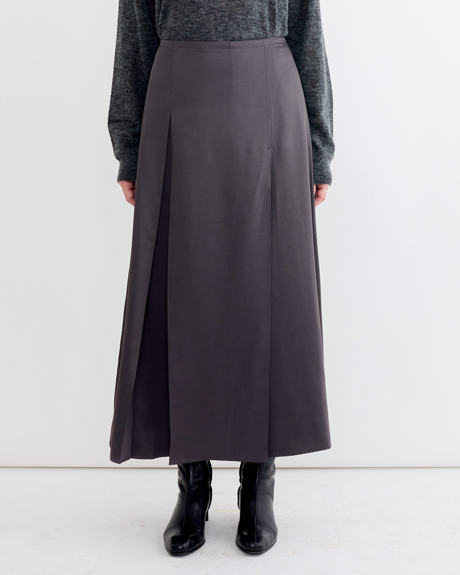 A person wearing the Studio Nicholson Youri Skirt in Slate, a long, high-waisted dark gray A-line skirt with subtle pleats, paired with a dark sweater and black ankle boots, stands against a plain white background.