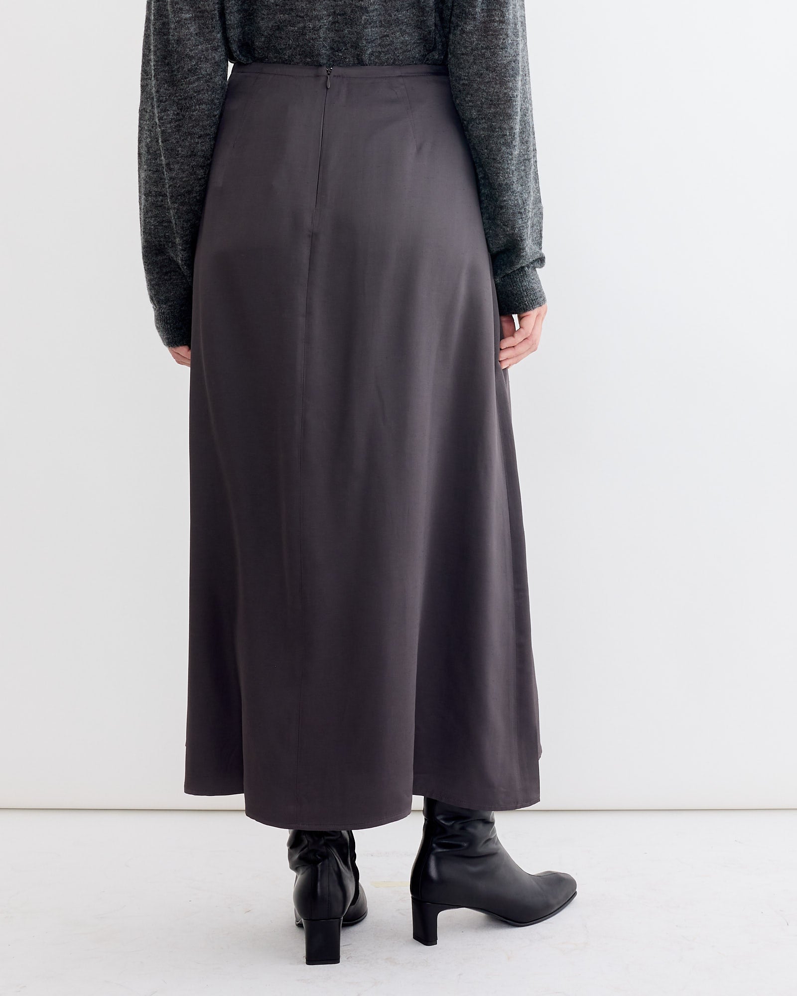 A person stands against a white background, wearing the Studio Nicholson Youri Skirt in Slate, paired with a matching sweater and black heeled boots. Photo is taken from behind with hands relaxed by their sides.