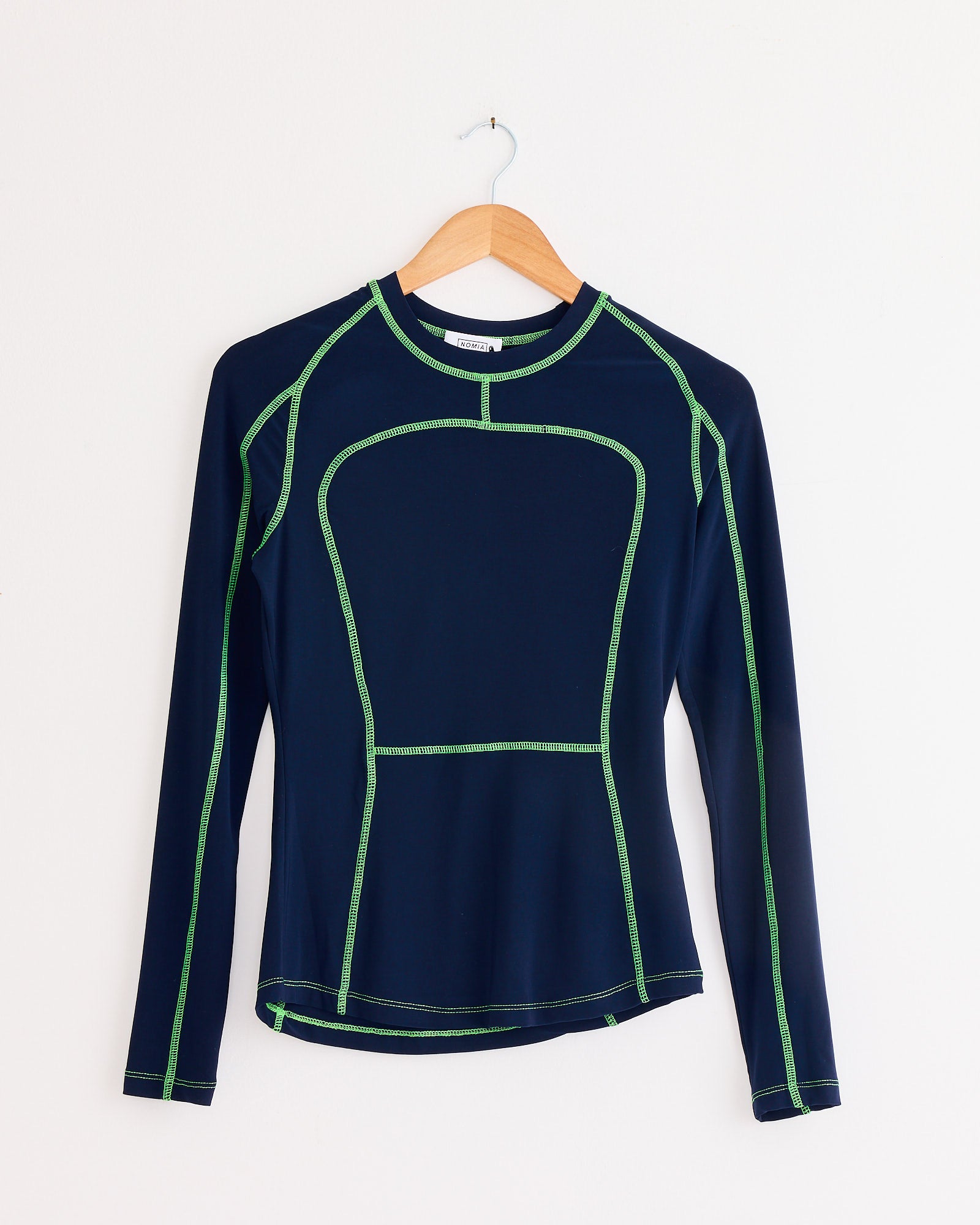 The NOMIA Paneled Scuba Top in Ink/Grass is a long-sleeve athletic top with bright green seams, made in New York, and shown on a wooden hanger against a plain white wall.