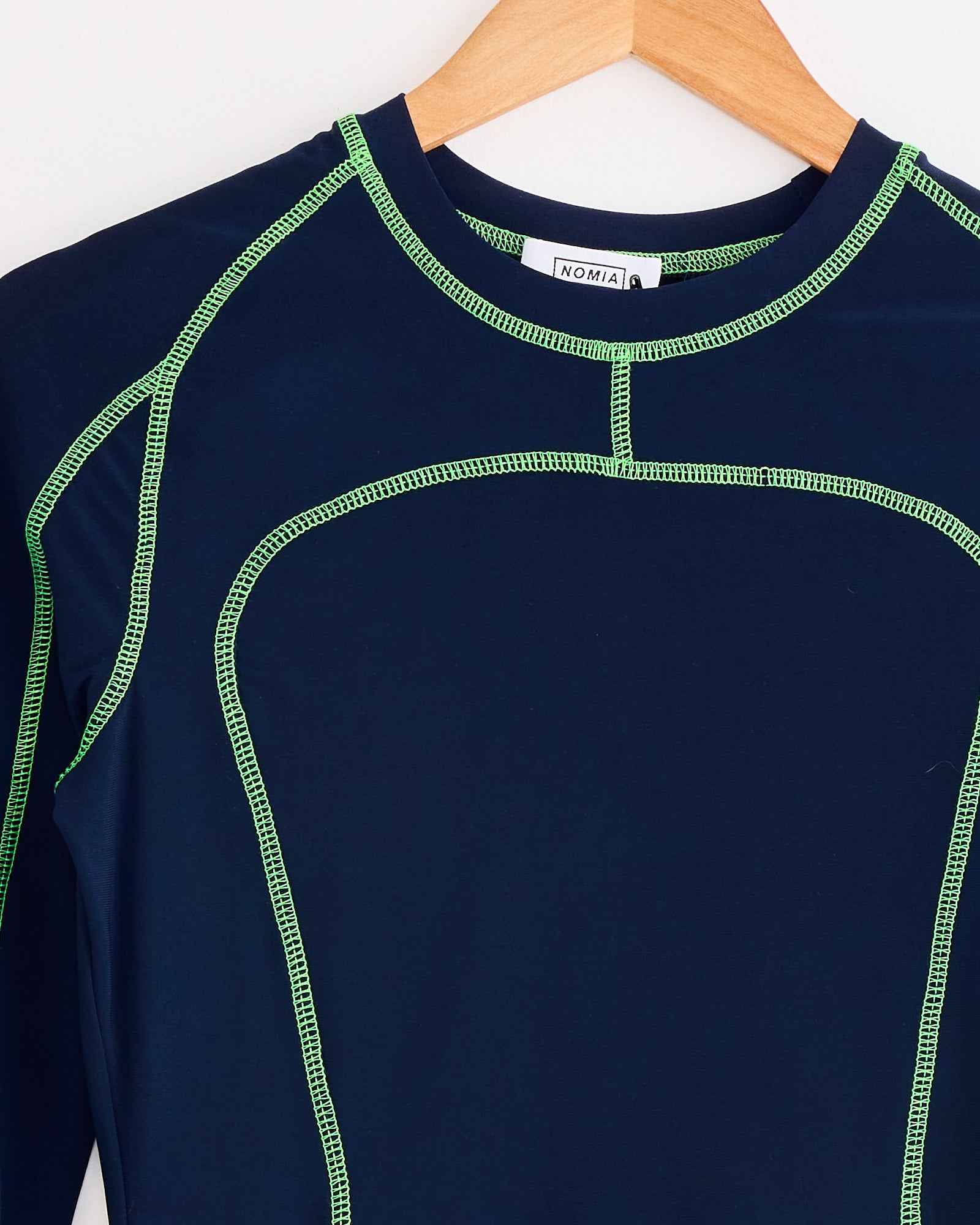 A close-up of the NOMIA Paneled Scuba Top in Ink / Grass, shown on a wooden hanger against a white background. Features include bright green contrast seams, a round neckline, and New York craftsmanship.