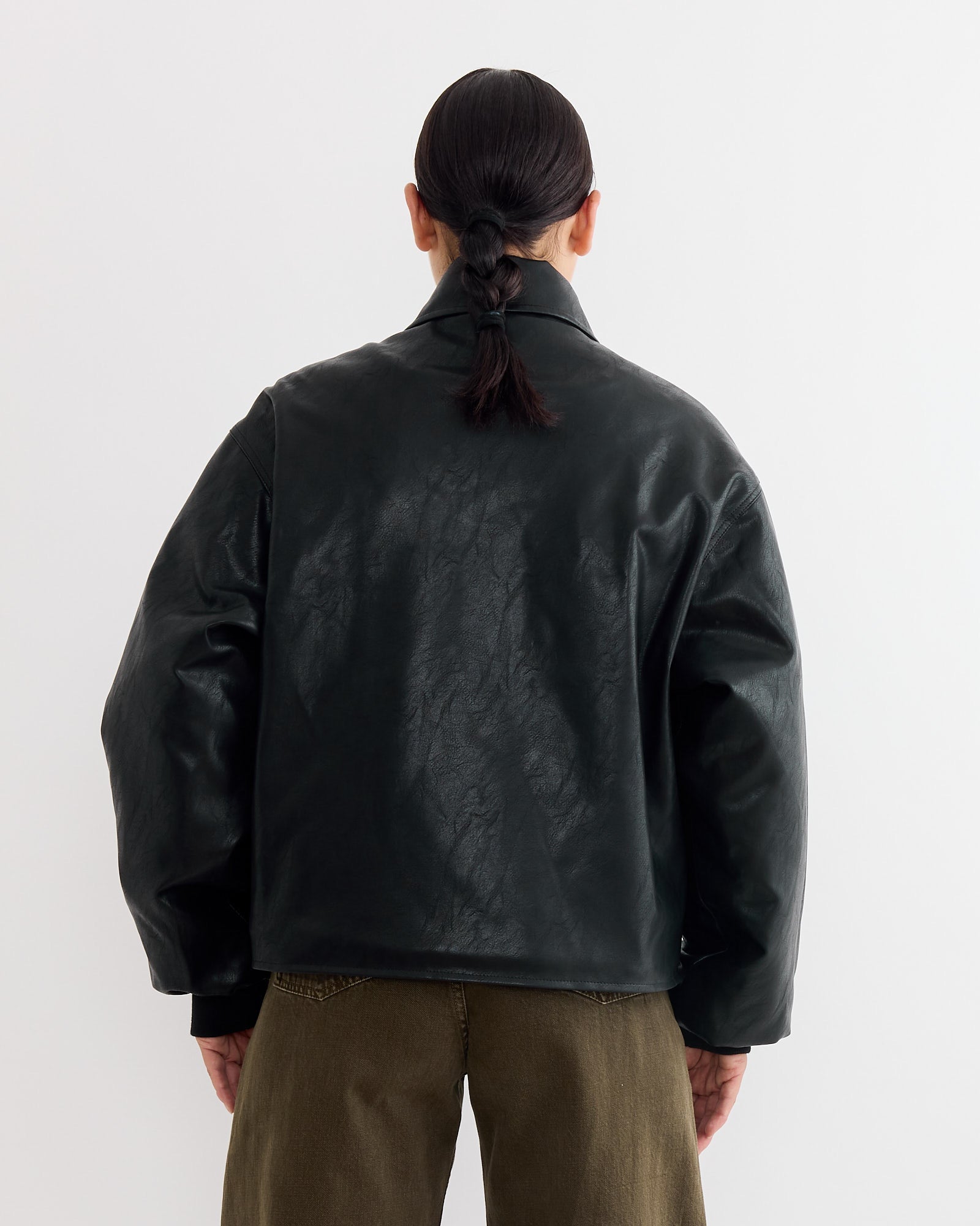 A person with a ponytail is wearing the NOMIA Faux Leather Bomber Jacket in Black.