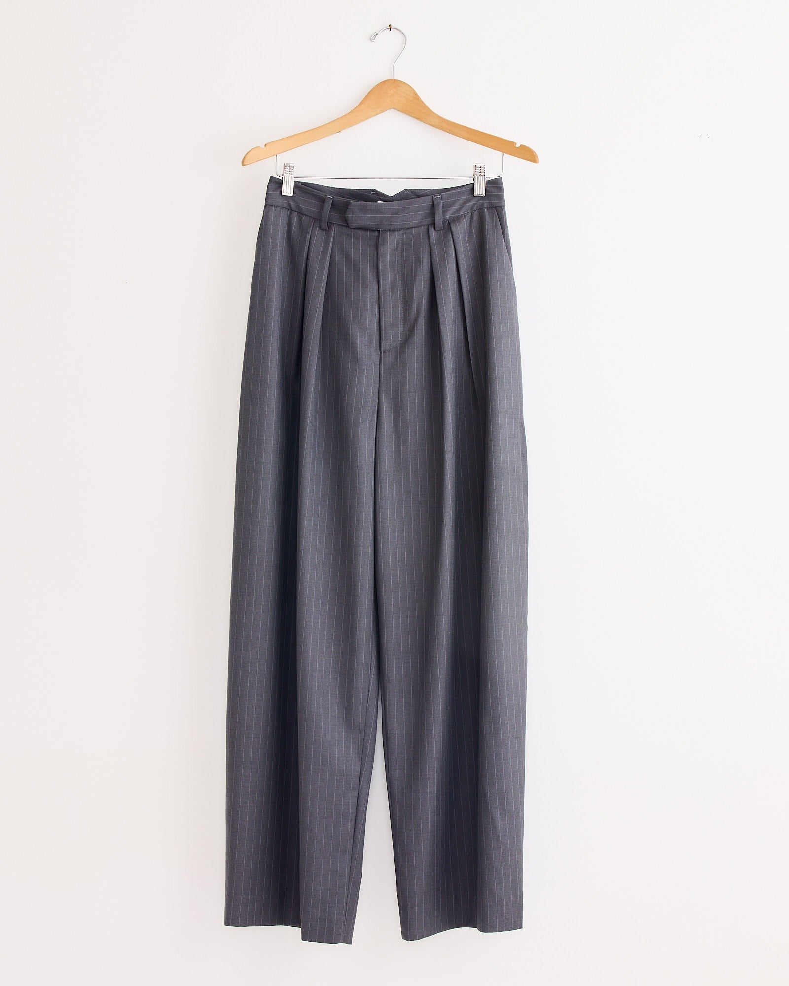 The NOMIA Wide Leg Pleated Trouser in Charcoal is a high-waisted, gray pinstripe twill pant made in NYC, shown hanging on a wooden hanger against a plain white wall.