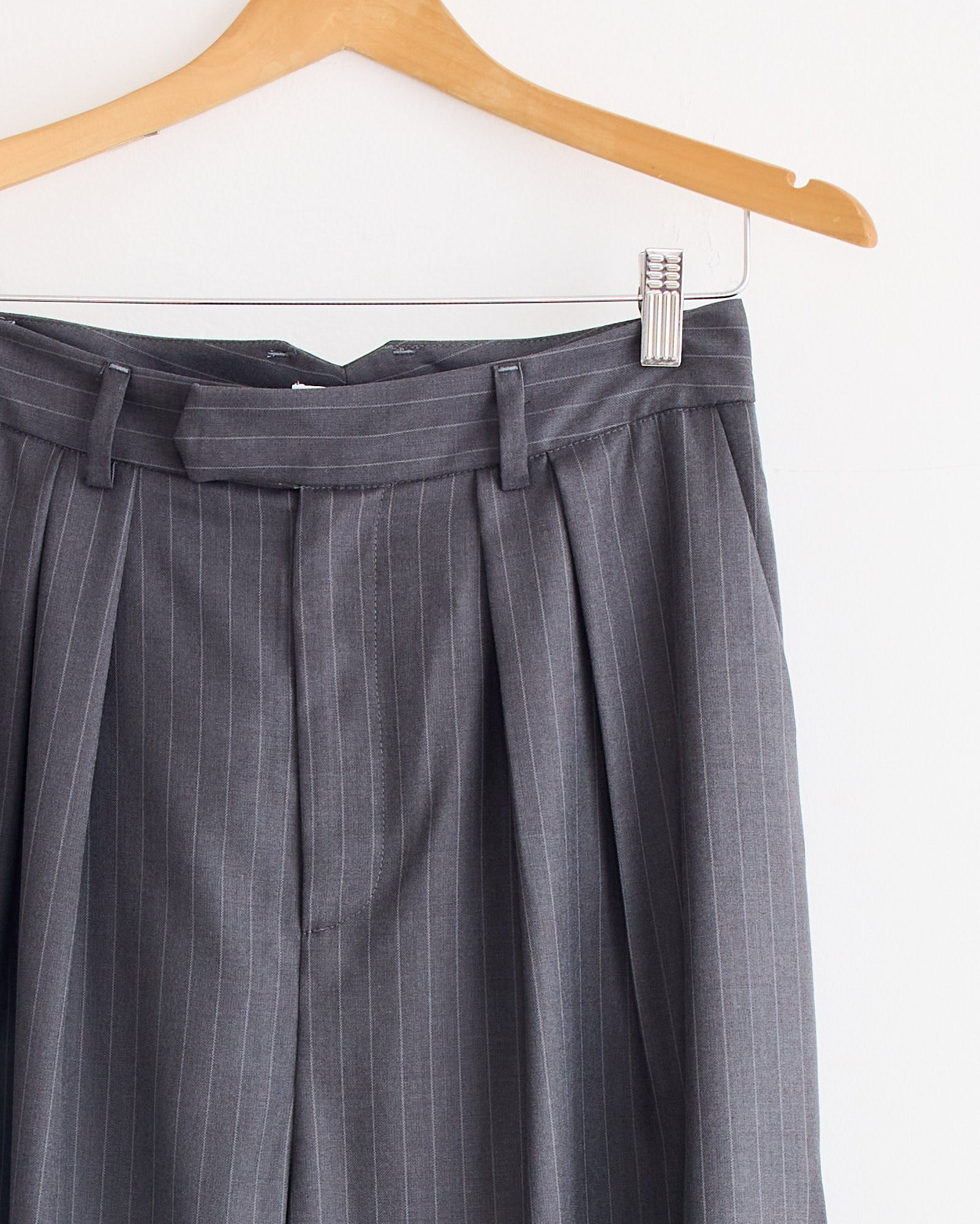 The upper half of the NOMIA Wide Leg Pleated Trouser in Charcoal is shown hanging on a wooden hanger against a white background, highlighting its pinstripe twill fabric, belt loops, and wide-leg pleated design.