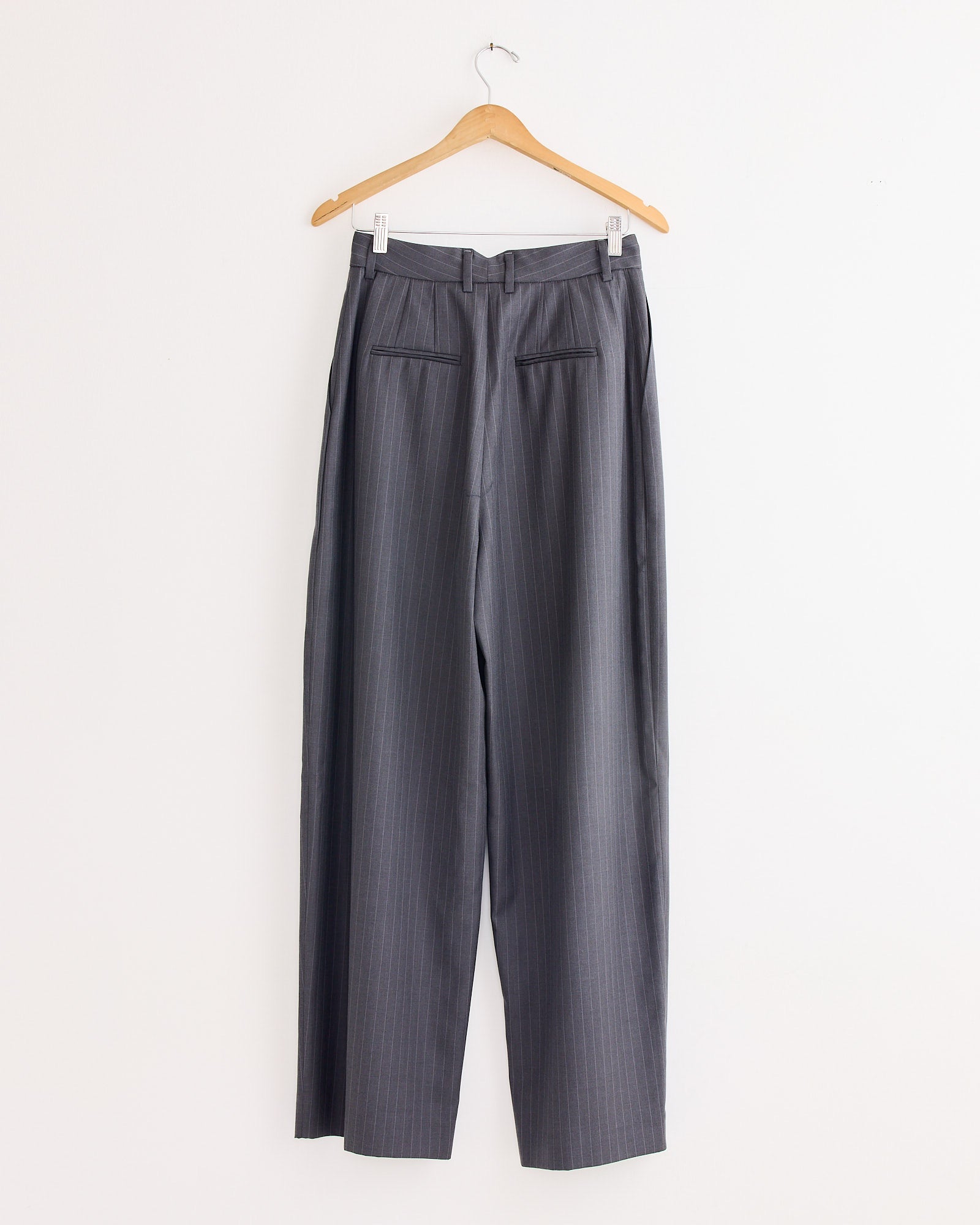 The Wide Leg Pleated Trouser in Charcoal by NOMIA hangs on a wooden hanger against a white wall, shown from the back to highlight two welt pockets. Made in NYC.