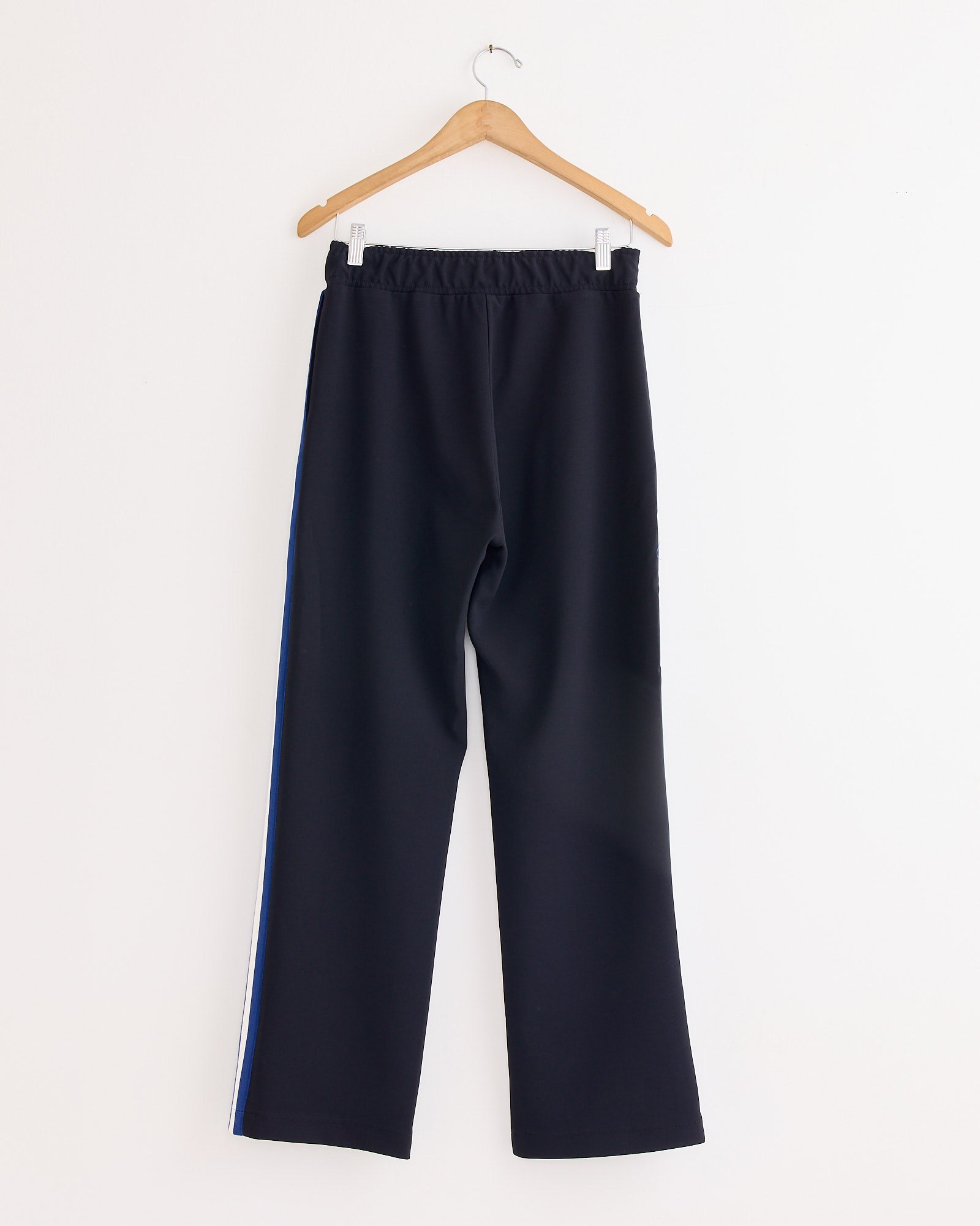The NOMIA Woven Stripe Track Pant in Black features blue and white side stripes, an elastic waistband, and is crafted from wrinkle-resistant crepe. Shown hanging on a wooden hanger against a plain white background.