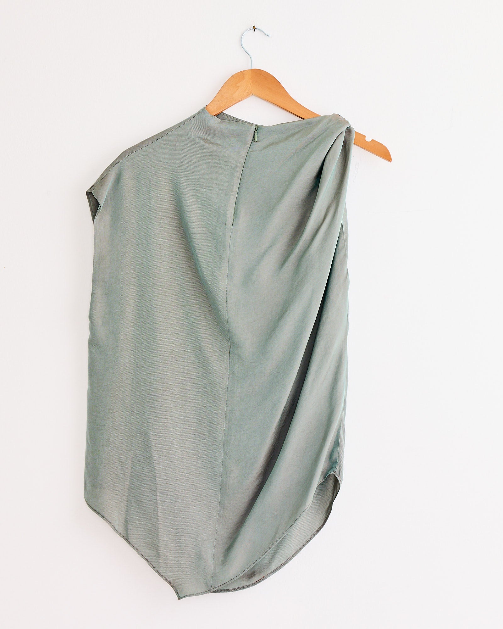 The NOMIA Twist Shoulder Top in Dusk is a light gray, asymmetrical sleeveless top with twisted shoulder detail, draped fabric, back zipper, and an uneven hemline. It's displayed on a wooden hanger against a white wall.