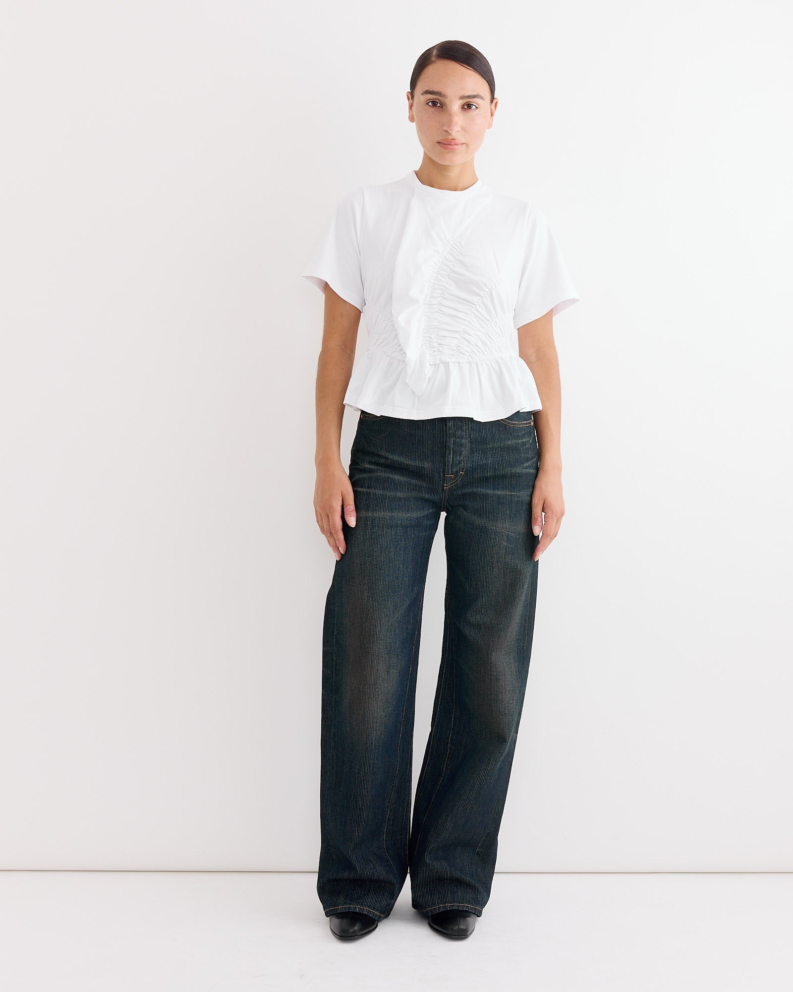 A person with dark hair pulled back and a neutral expression stands against a white wall, wearing the Cecilie Bahnsen Ace T-Shirt in White with ruffle detail, paired with dark wide-leg jeans and black shoes.