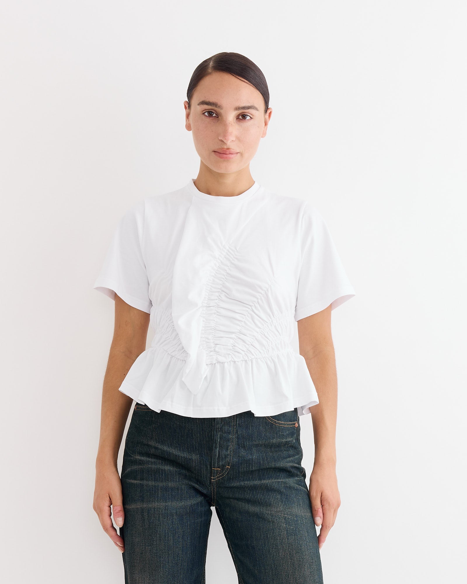 A woman with dark hair pulled back models the Cecilie Bahnsen Ace T-Shirt in White, a short-sleeve blouse with ruffle detail and peplum hem, paired with dark denim jeans, against a plain white background.