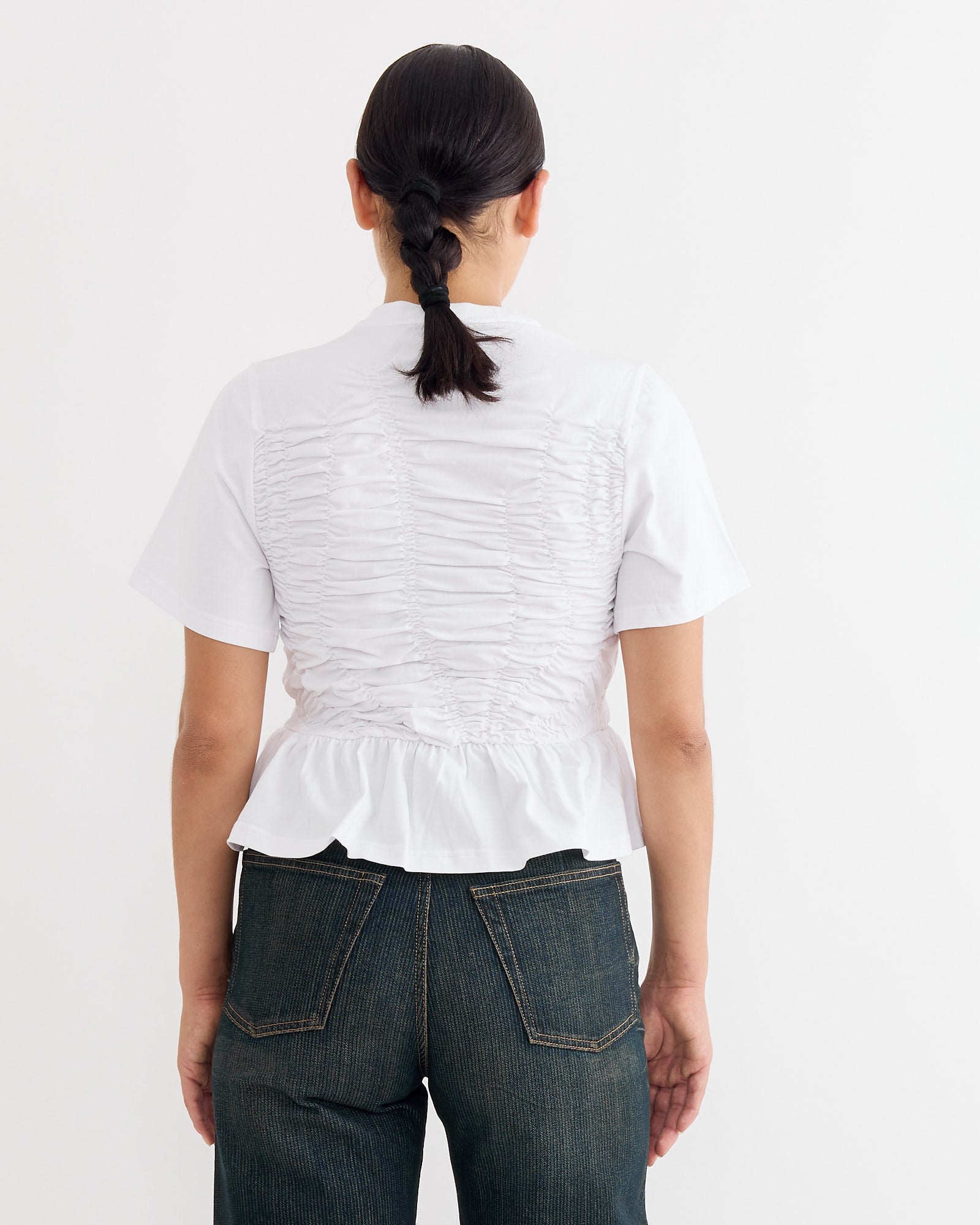A person with dark braided hair wears the Cecilie Bahnsen Ace T-Shirt in White, featuring ruched back detailing, paired with dark blue jeans. They stand facing away from the camera against a plain light background.
