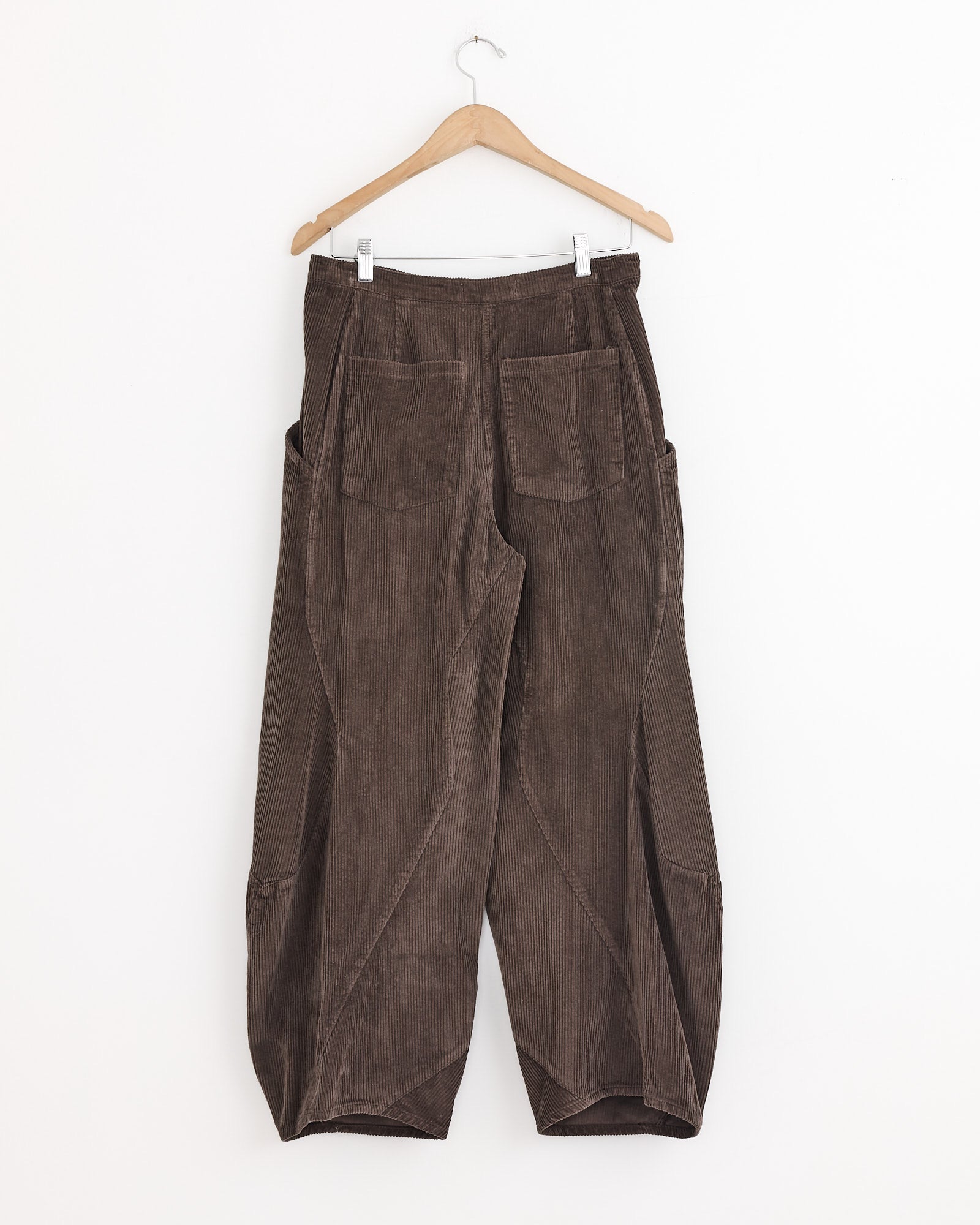 The Henrik Vibskov Digi Corduroy Pant in Chocolate Brown, featuring wide legs and large back pockets, hangs on a wooden hanger against a plain white wall.