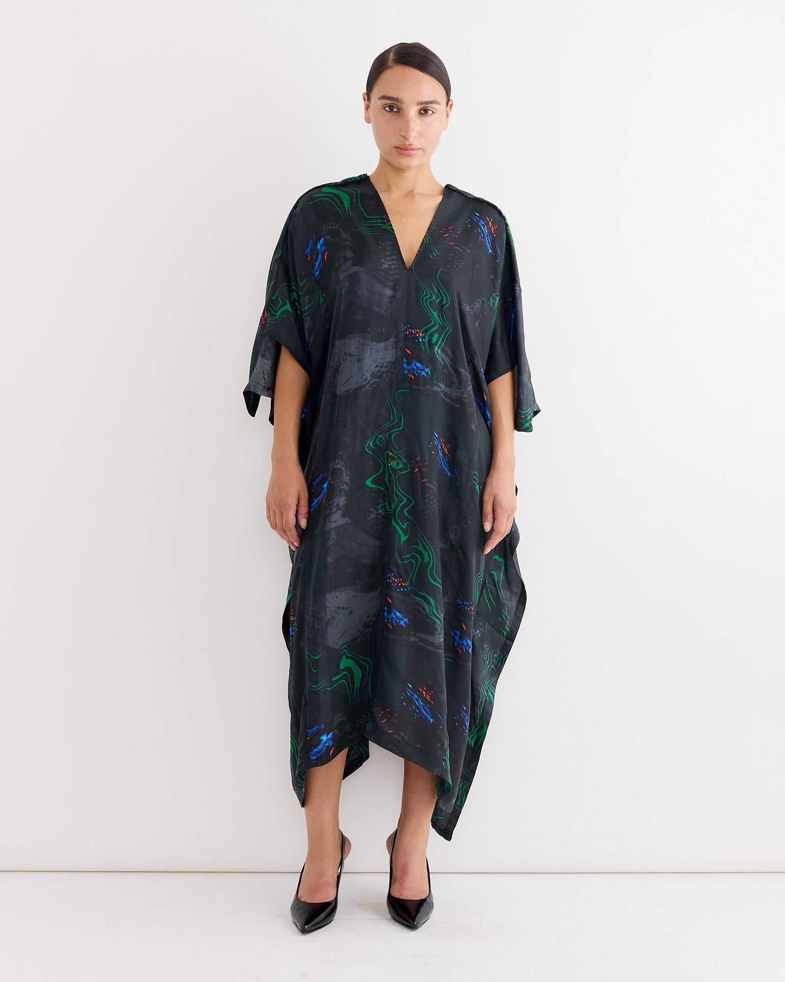 A woman stands against a white wall, wearing Henrik Vibskov's Uncut Kaftan in Dark Fireworks—a loose, dark dress with a bold multicolour print. Her straight, dark hair is pulled back and she completes the look with black pointed-toe heels.