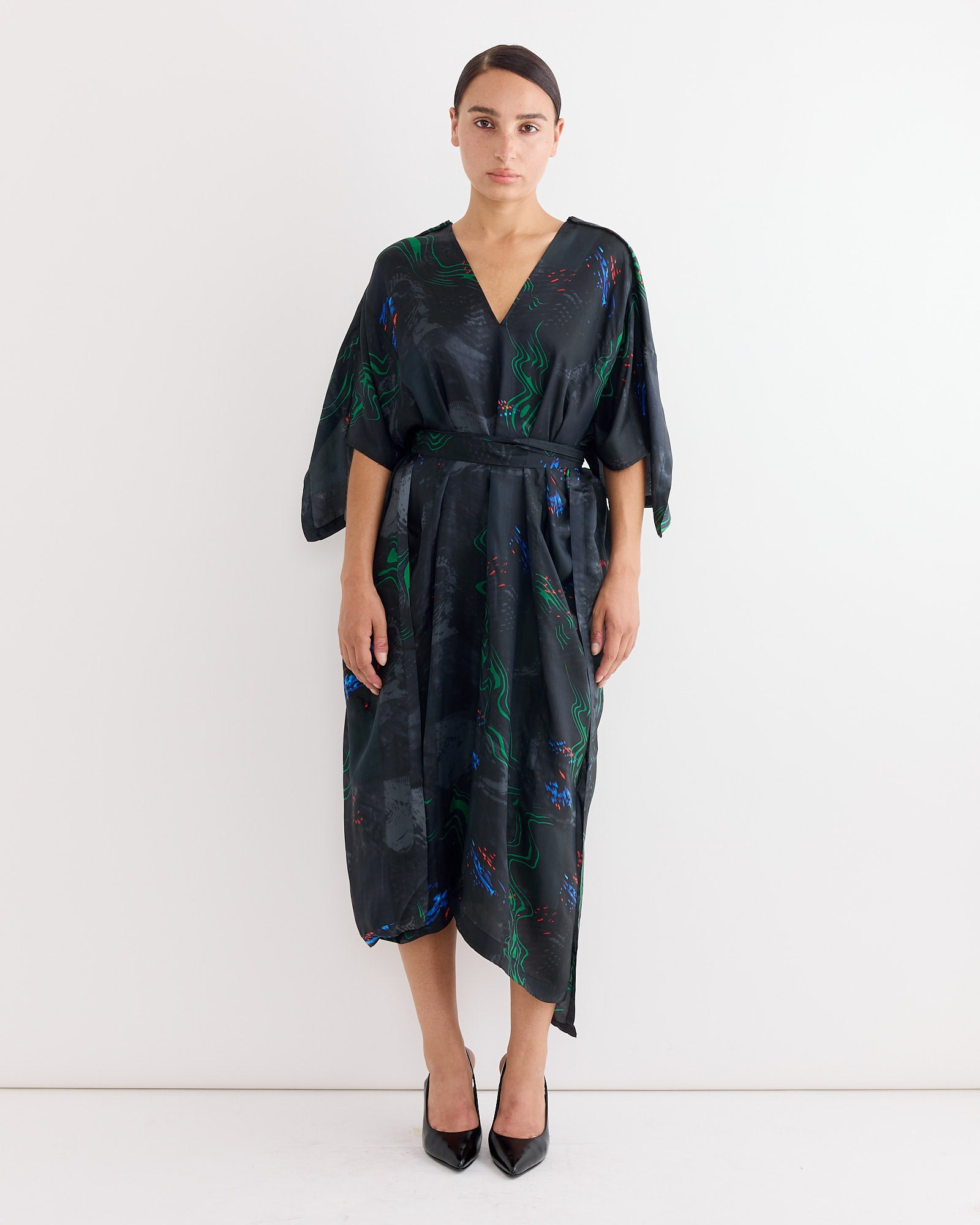 A woman with her hair pulled back stands against a white wall wearing the Henrik Vibskov Uncut Kaftan in Dark Fireworks, a loose, knee-length multicolour print dress with a V-neck, paired with black pointed-toe heels.