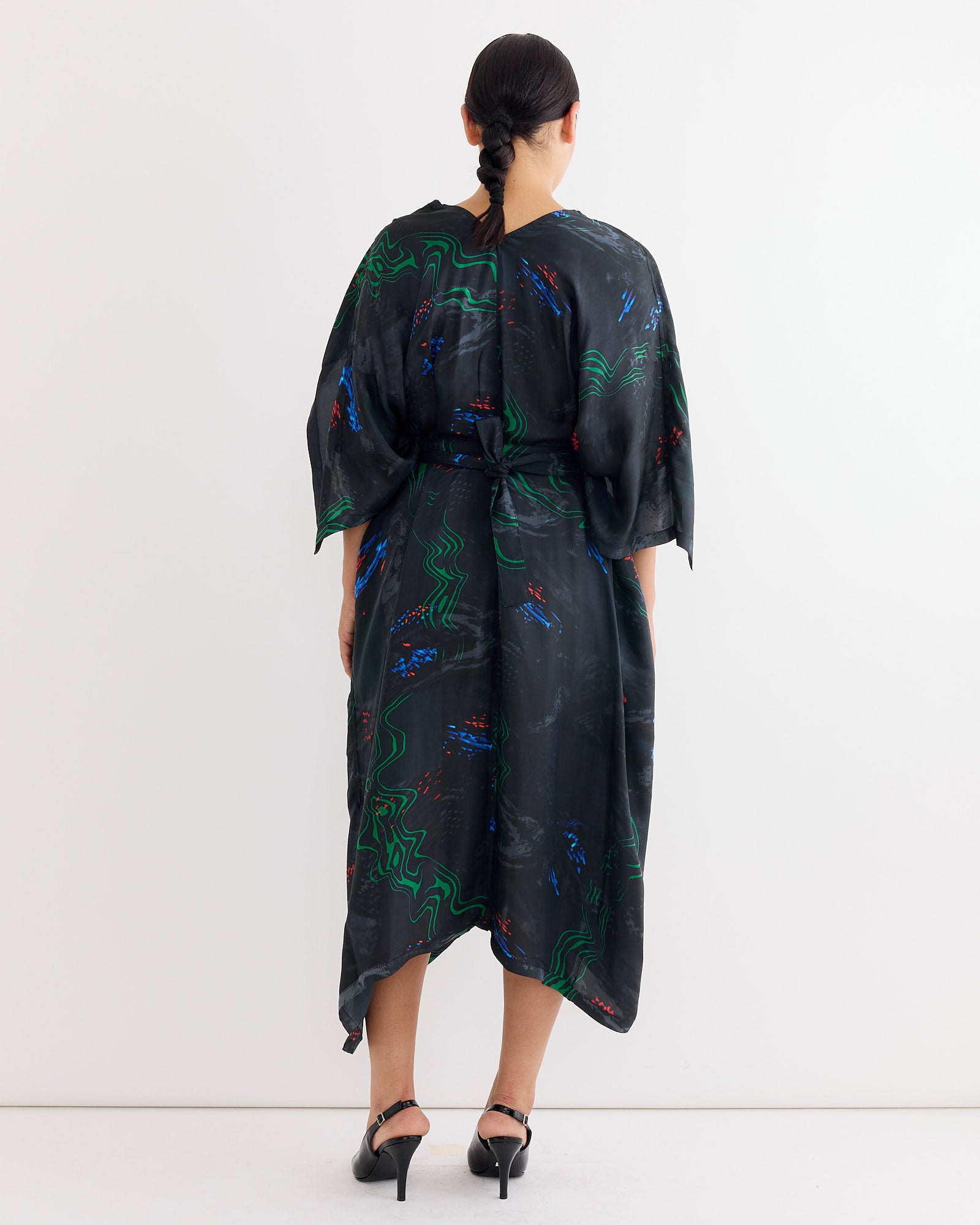 A person with dark braided hair faces away, wearing the Henrik Vibskov Uncut Kaftan in Dark Fireworks with a multicolour print and asymmetrical hem, paired with black heeled sandals, set against a plain white background.