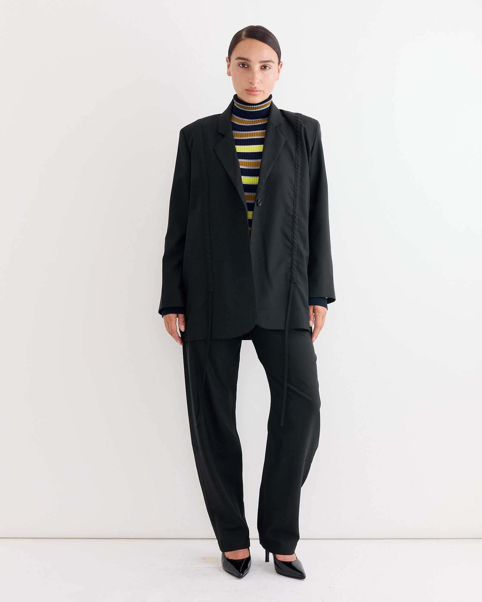 A person stands against a white wall, wearing the Henrik Vibskov Backpack Blazer in Tropical Black, paired with black dress pants, a striped yellow, black, and white turtleneck sweater, and black pointed-toe heels.