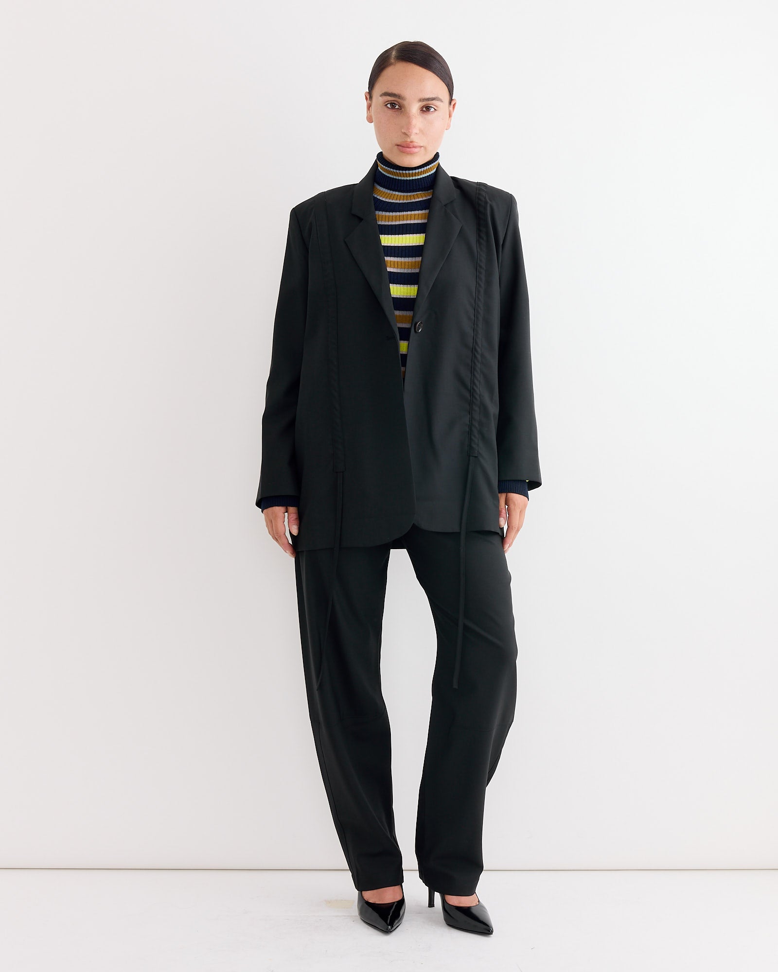 A person stands against a white background wearing the Henrik Vibskov Banana Pant in Tropical Black, styled with a loose black suit, black heels, and a black, yellow, and white striped turtleneck. Their hair is pulled back and arms rest at their sides.