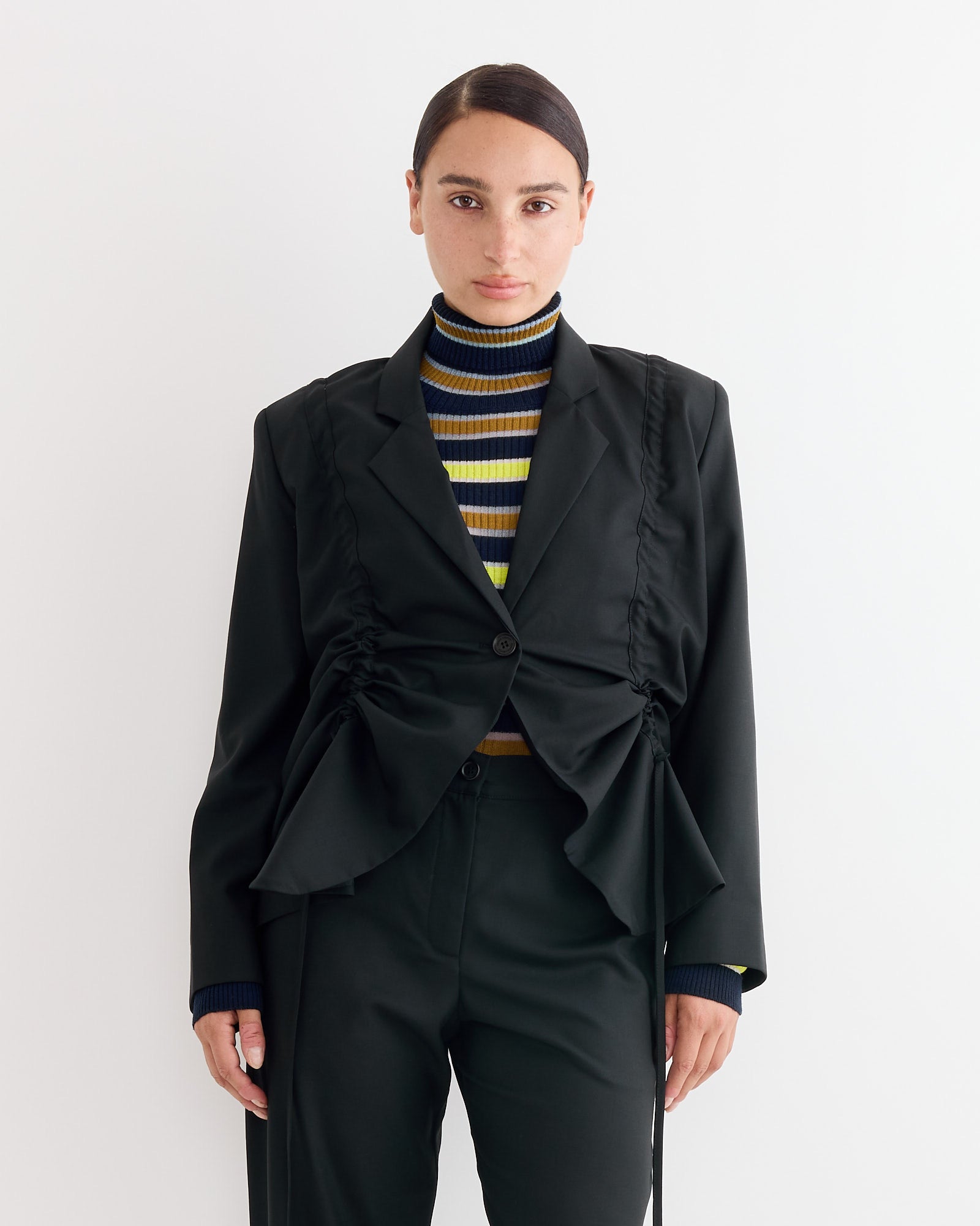 A person with dark hair pulled back wears the Henrik Vibskov Backpack Blazer in Tropical Black over a striped turtleneck with yellow, blue, black, and white stripes, standing against a plain white background.
