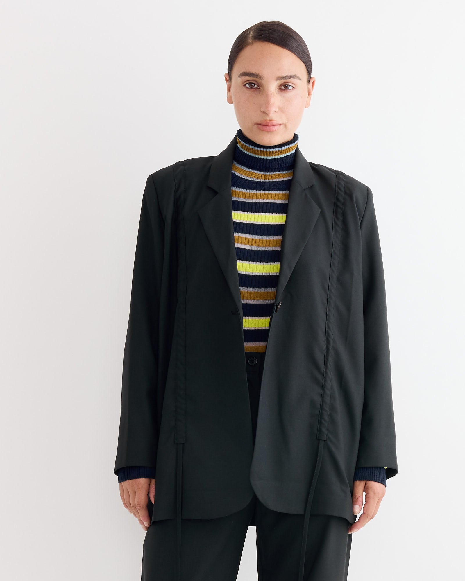 A person with dark hair pulled back wears the Henrik Vibskov Backpack Blazer in Tropical Black over a striped turtleneck sweater, standing against a plain light background.