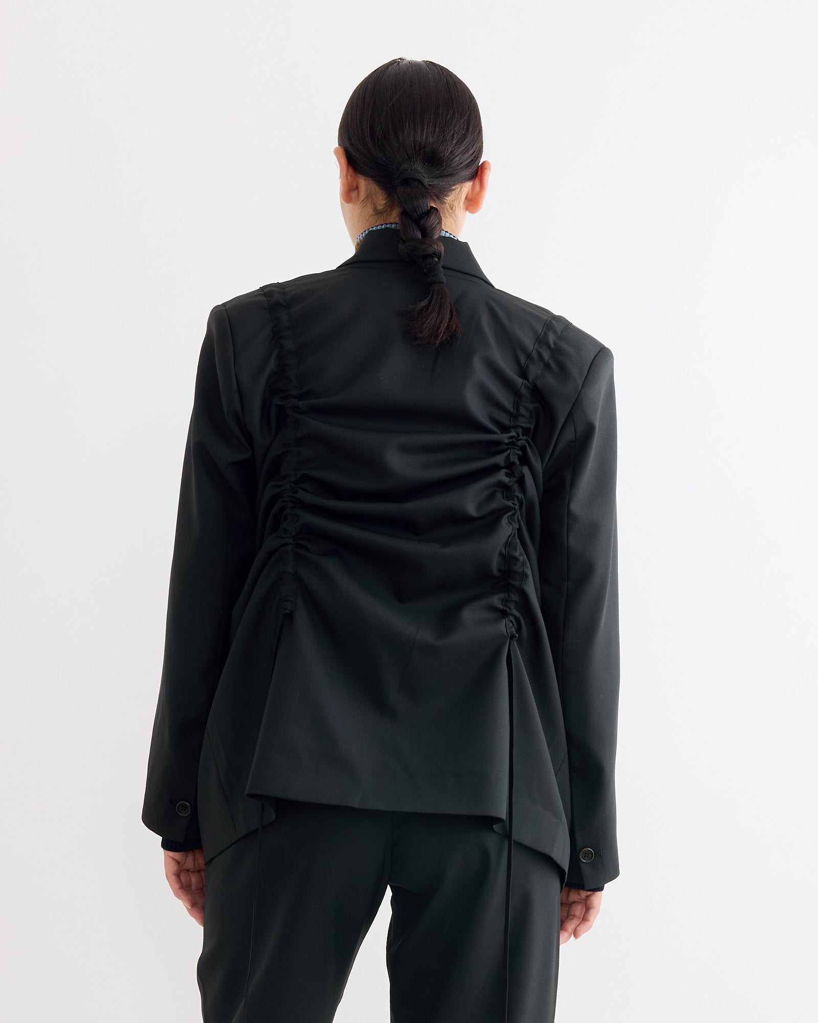 Seen from behind, a person with dark hair in a braided ponytail wears the Henrik Vibskov Backpack Blazer in Tropical Black with matching trousers, standing against a plain white background.