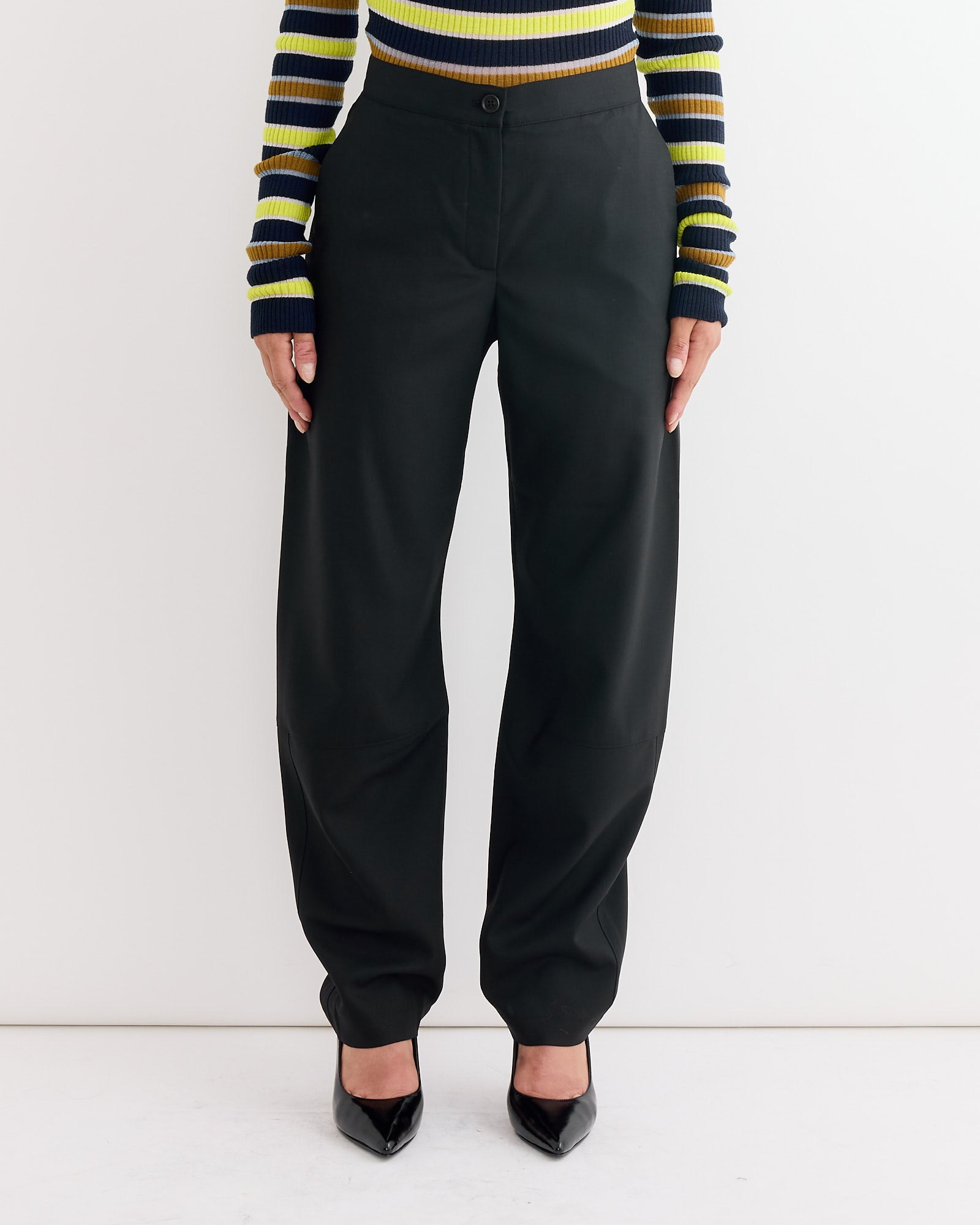 A person wears Henrik Vibskov's Banana Pant in Tropical Black (high-waisted, loose fit, 100% wool), a yellow-blue-black striped long-sleeve top, and black pointed-toe heels against a plain white background.