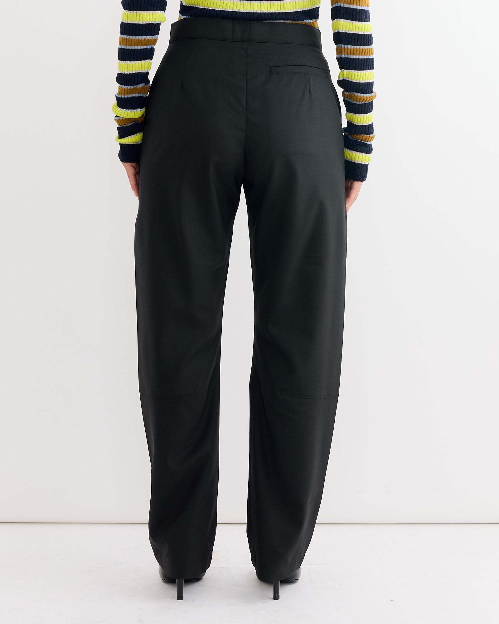 Person stands with their back to the camera, wearing Henrik Vibskov's Banana Pant in Tropical Black, black heeled shoes, and a ribbed long-sleeve top featuring yellow, green, and black horizontal stripes.