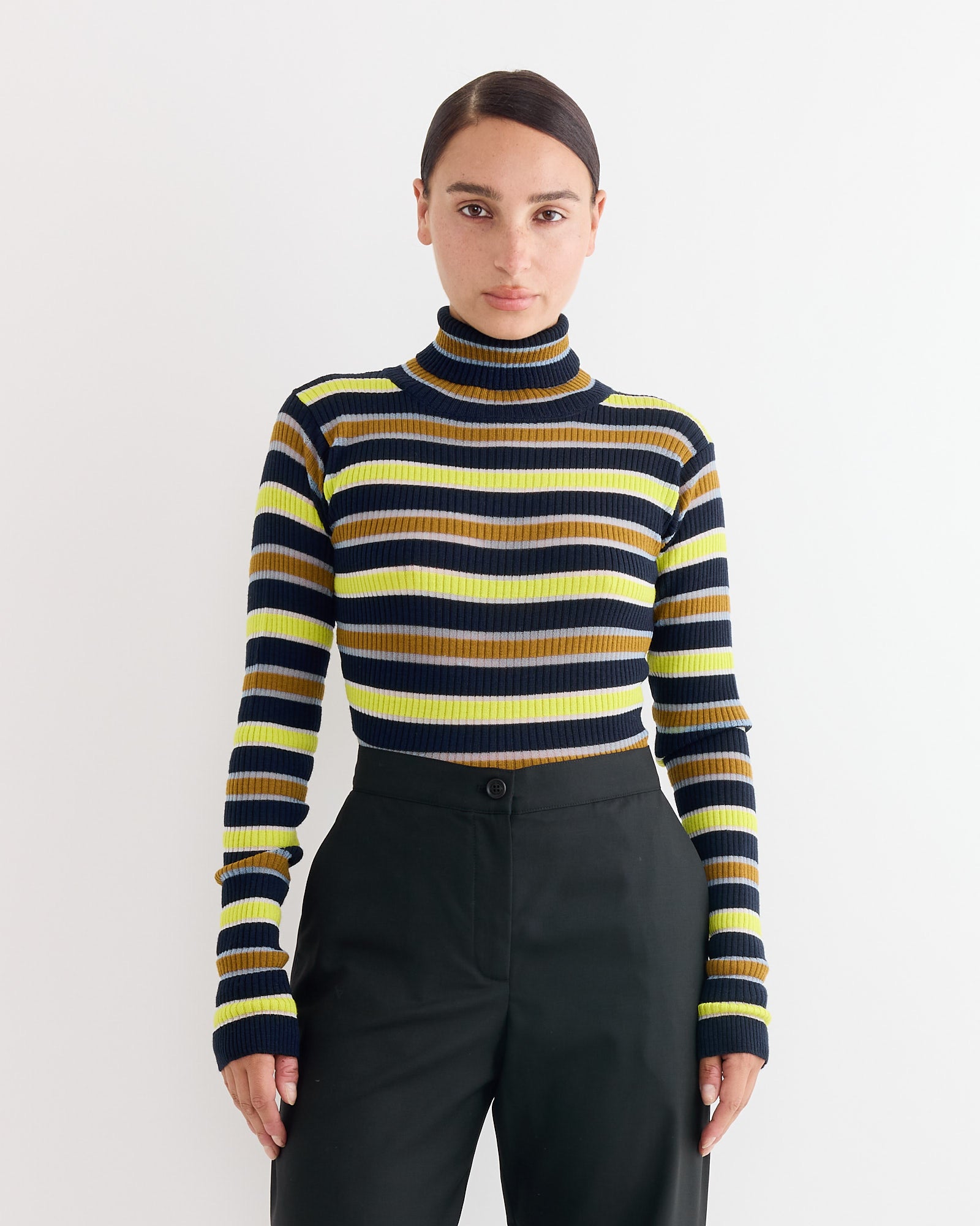 A person with dark, center-parted hair pulled back wears the Henrik Vibskov Line Turtleneck in Navy Stripes with horizontal yellow, black, brown, and navy lines, paired with high-waisted black pants against a plain white background.