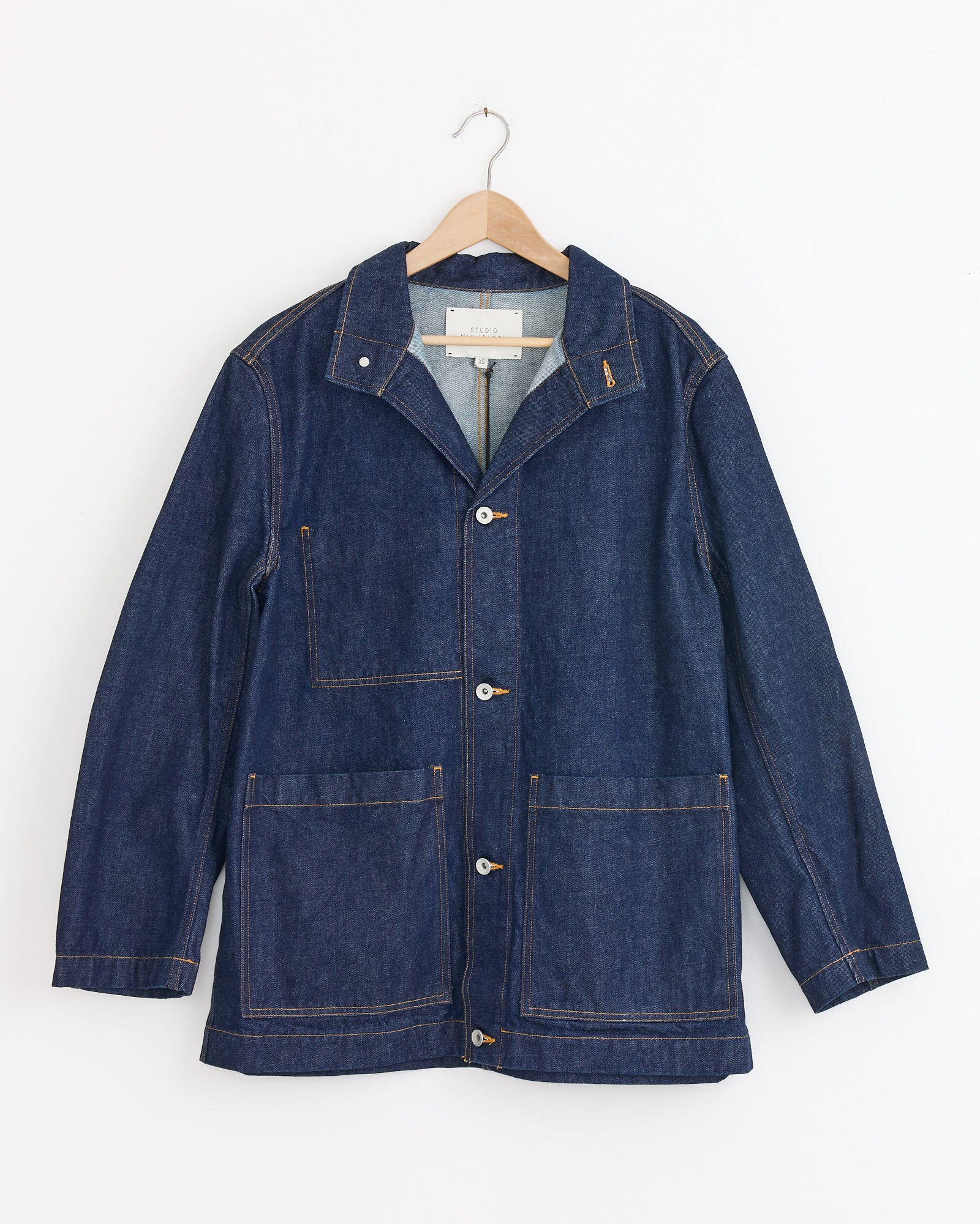 The Studio Nicholson District Denim Jacket in Indigo Wash, inspired by classic workwear with large front pockets, button closure, and brown stitching, hangs on a wooden hanger against a white background.