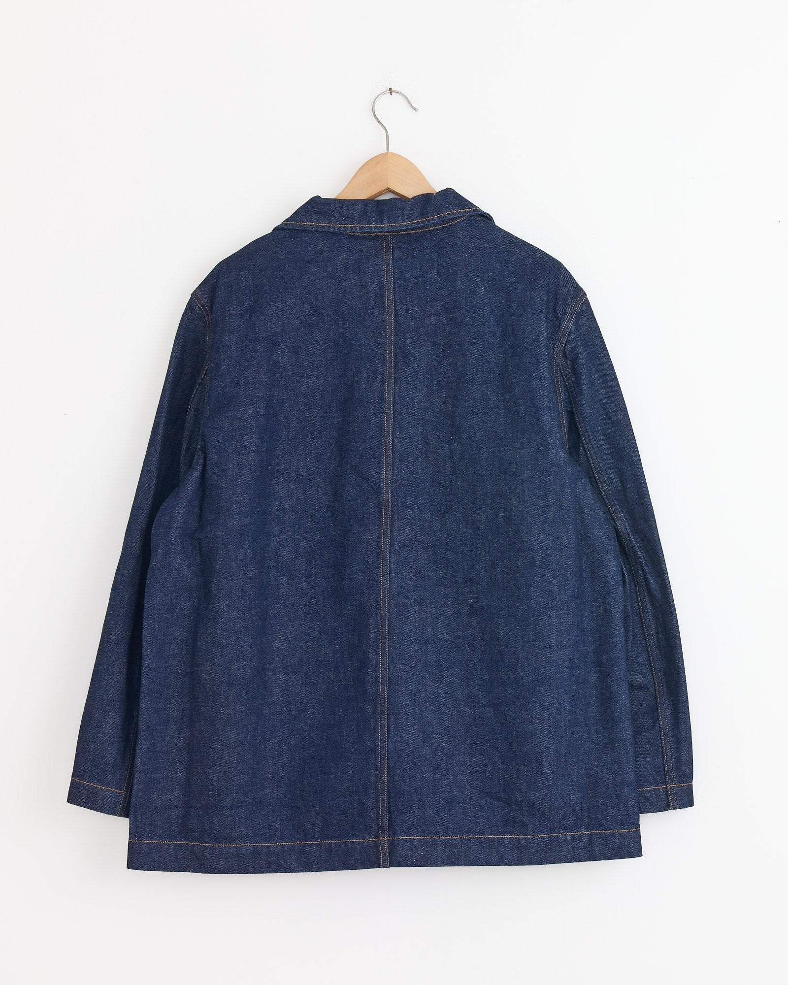 The Studio Nicholson District Denim Jacket in Indigo Wash hangs on a wooden hanger against a white wall, shown from the back with visible stitching and a minimalist, collarless design inspired by classic Japanese workwear.