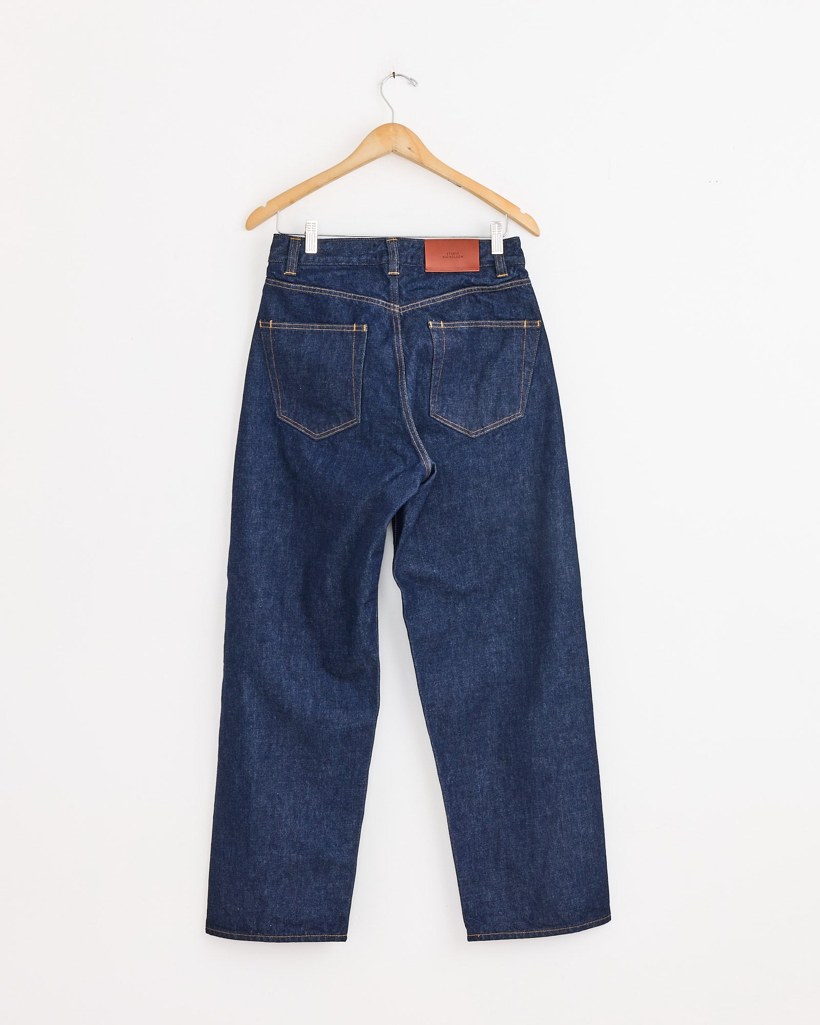 The Seb Jean in Indigo Wash by Studio Nicholson hangs on a wooden hanger against a white wall, displayed from the back with visible rear pockets and a brown leather patch on the waistband.