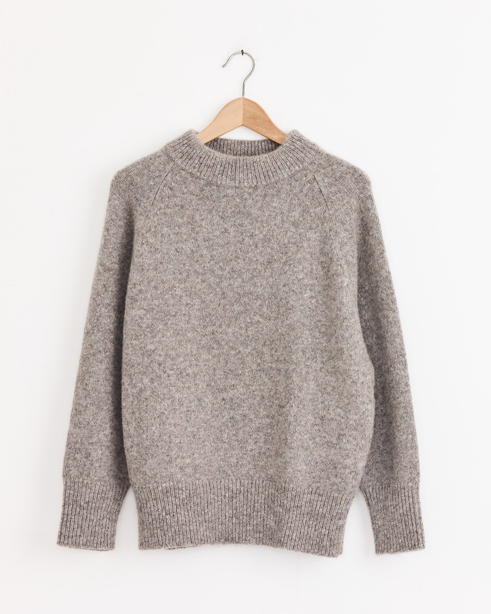 A Studio Nicholson Persephone Crewneck in Natural, crafted from baby alpaca knit with long sleeves and ribbed trim, hangs on a wooden hanger against a plain white wall.
