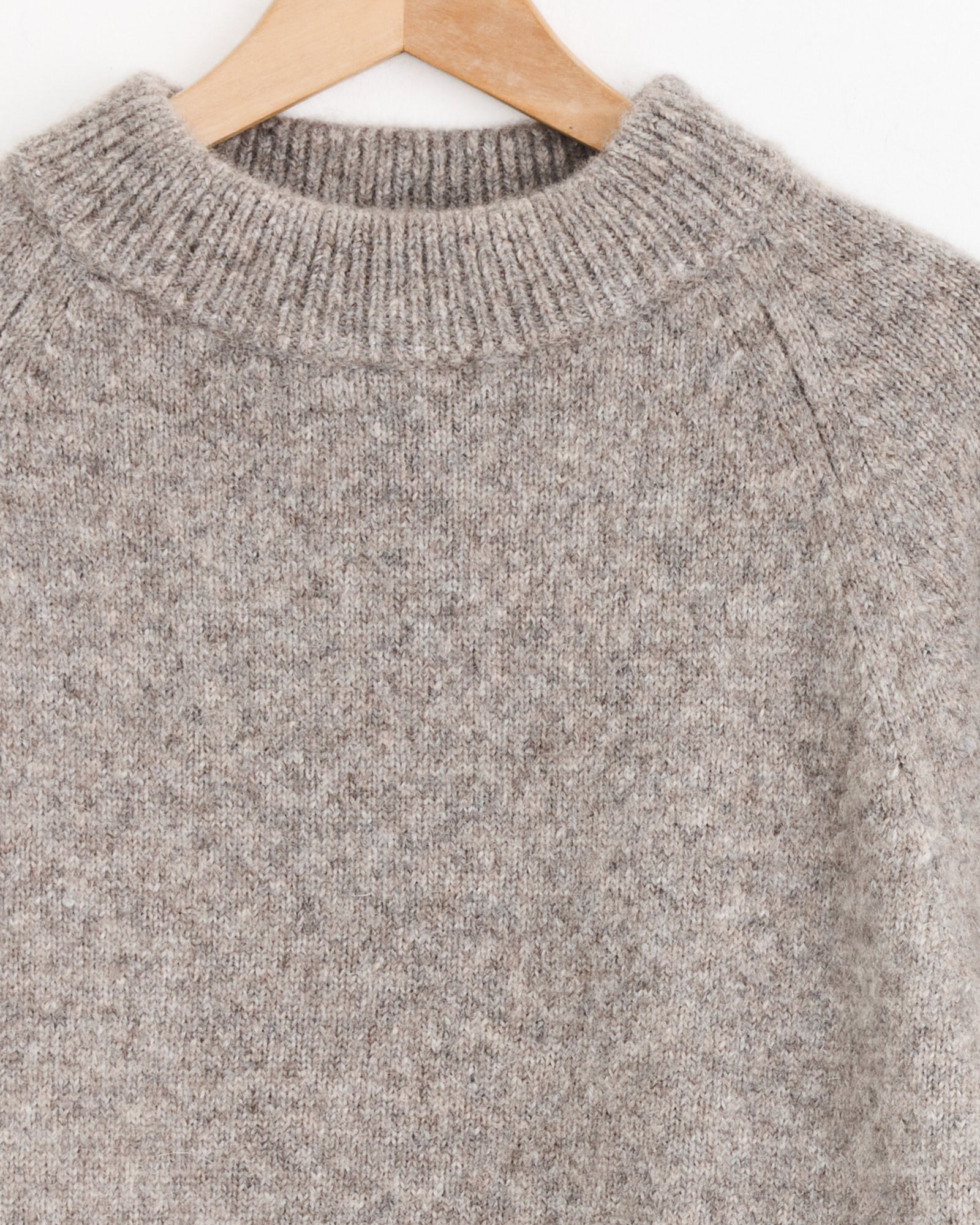 A close-up of the Studio Nicholson Persephone Crewneck in Natural, featuring a light brown ribbed collar, hangs on a wooden hanger against a plain white background.